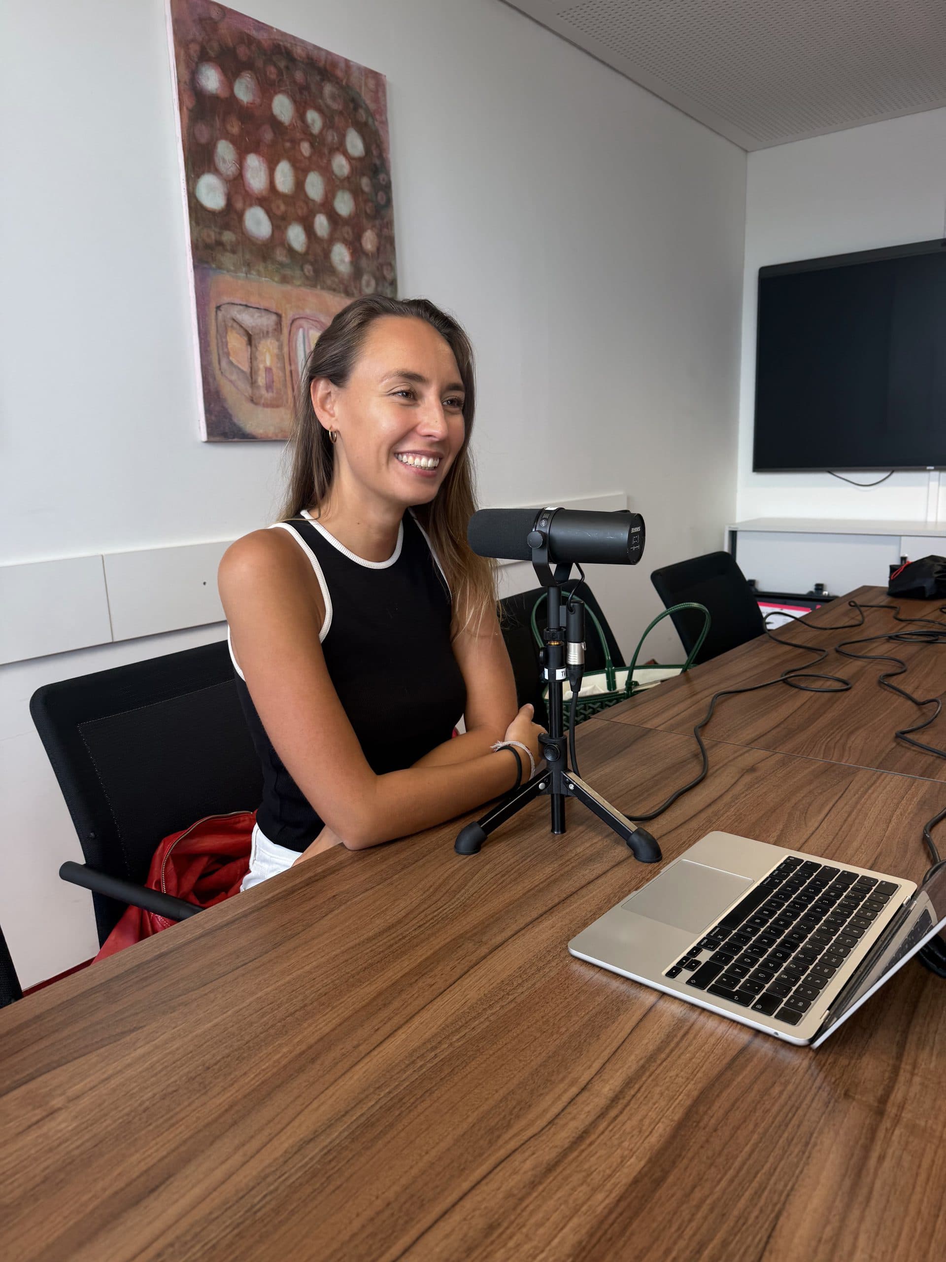 Eine Frau sitzt an einem Konferenztisch, lächelt und spricht in ein Mikrofon, vor ihr steht ein Laptop. Sie scheint einen Podcast oder ein Interview in einer modernen Büroumgebung aufzunehmen.