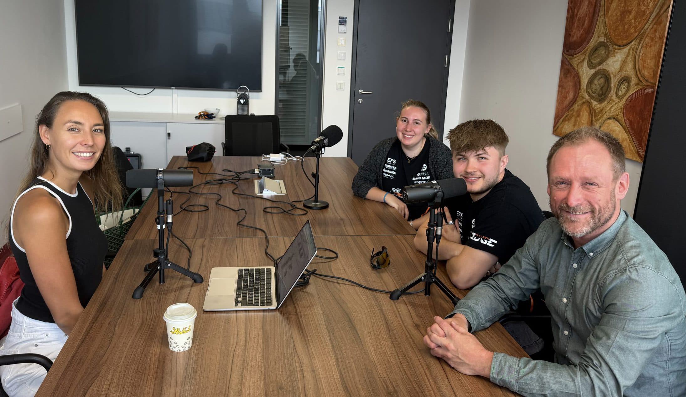 Vier Personen sitzen mit Mikrofonen und einem Laptop um einen Tisch und scheinen bereit zu sein, einen Podcast aufzunehmen. Auf dem Tisch stehen Kaffeetassen und Kabel. Im Hintergrund sind ein großer Bildschirm und ein Kunstwerk zu sehen.
