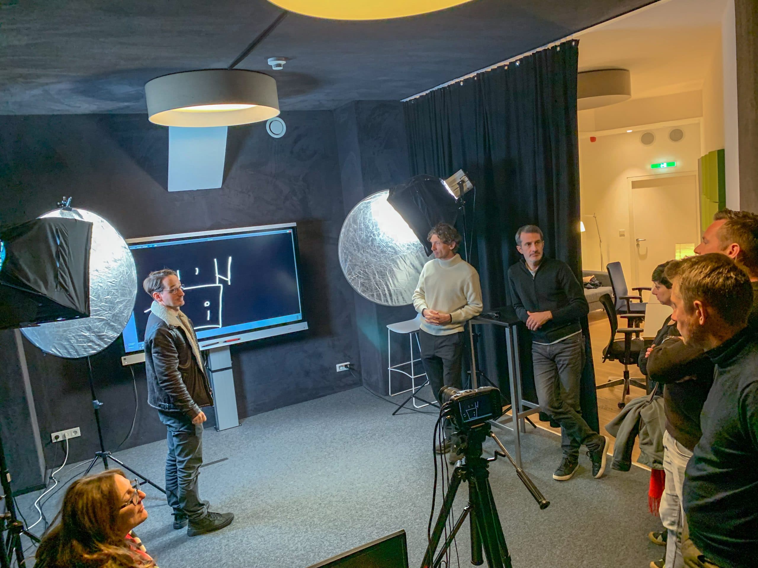 Fünf Personen stehen in einem Studio mit professioneller Beleuchtung und Kameras, konzentriert auf einen Mann, der vor einem Bildschirm mit einfachen weißen Diagrammen präsentiert. Der Raum ist mit dunklen Wänden und modernen Beleuchtungskörpern ausgestattet.