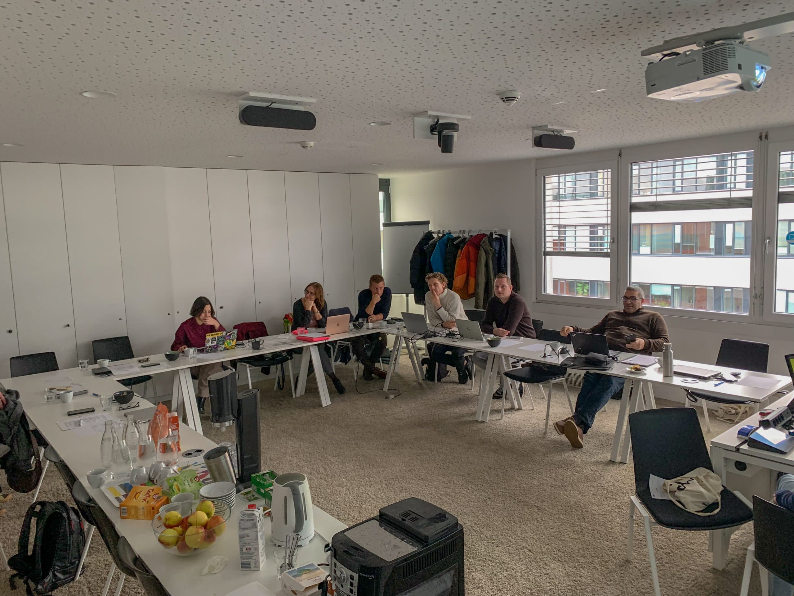 In einem hellen Besprechungsraum sitzt eine Gruppe von Menschen an U-förmig angeordneten Tischen, auf denen Laptops, Papiere und Snacks stehen. Im hinteren Bereich hängen Mäntel, und große Fenster zeigen Gebäude im Freien.