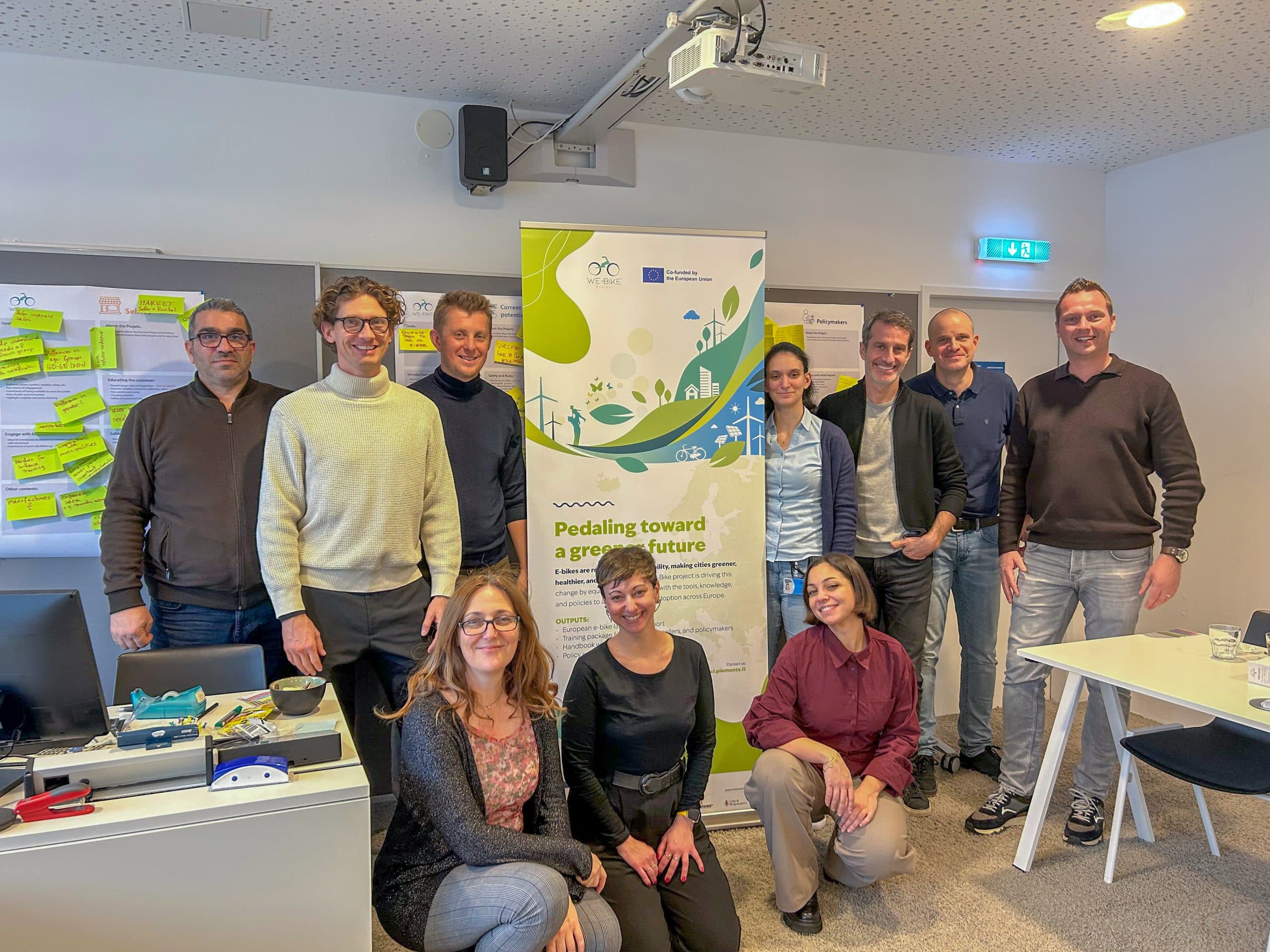 Zehn Menschen posieren drinnen um ein Transparent mit der Aufschrift 'Pedalling towards a greener future'. Einige stehen, einige knien. Im Hintergrund des Raums befinden sich ein Whiteboard, Haftnotizen und Bürogeräte.