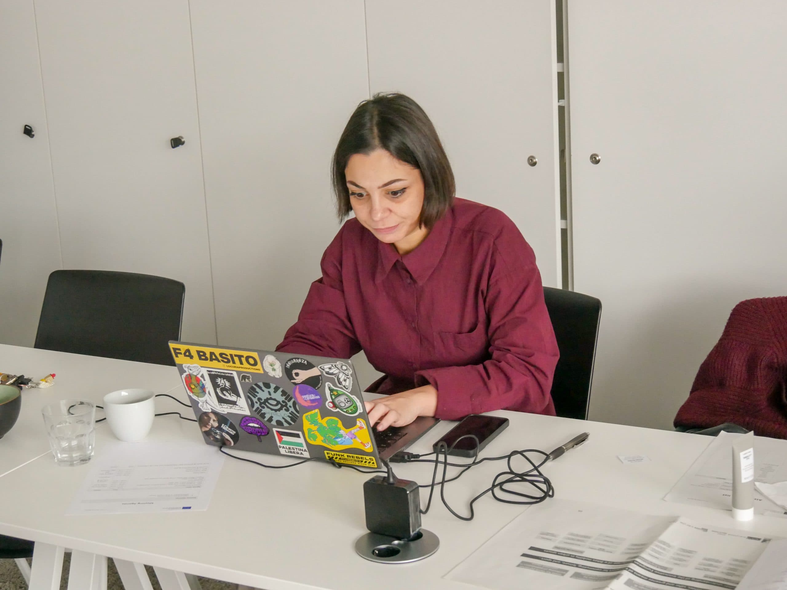 Eine Frau in einem kastanienbraunen Hemd arbeitet an einem mit Aufklebern versehenen Laptop an einem weißen Schreibtisch mit Papieren, einem Telefon, einer Tasse und Kabeln um sie herum in einer modernen Büroumgebung.