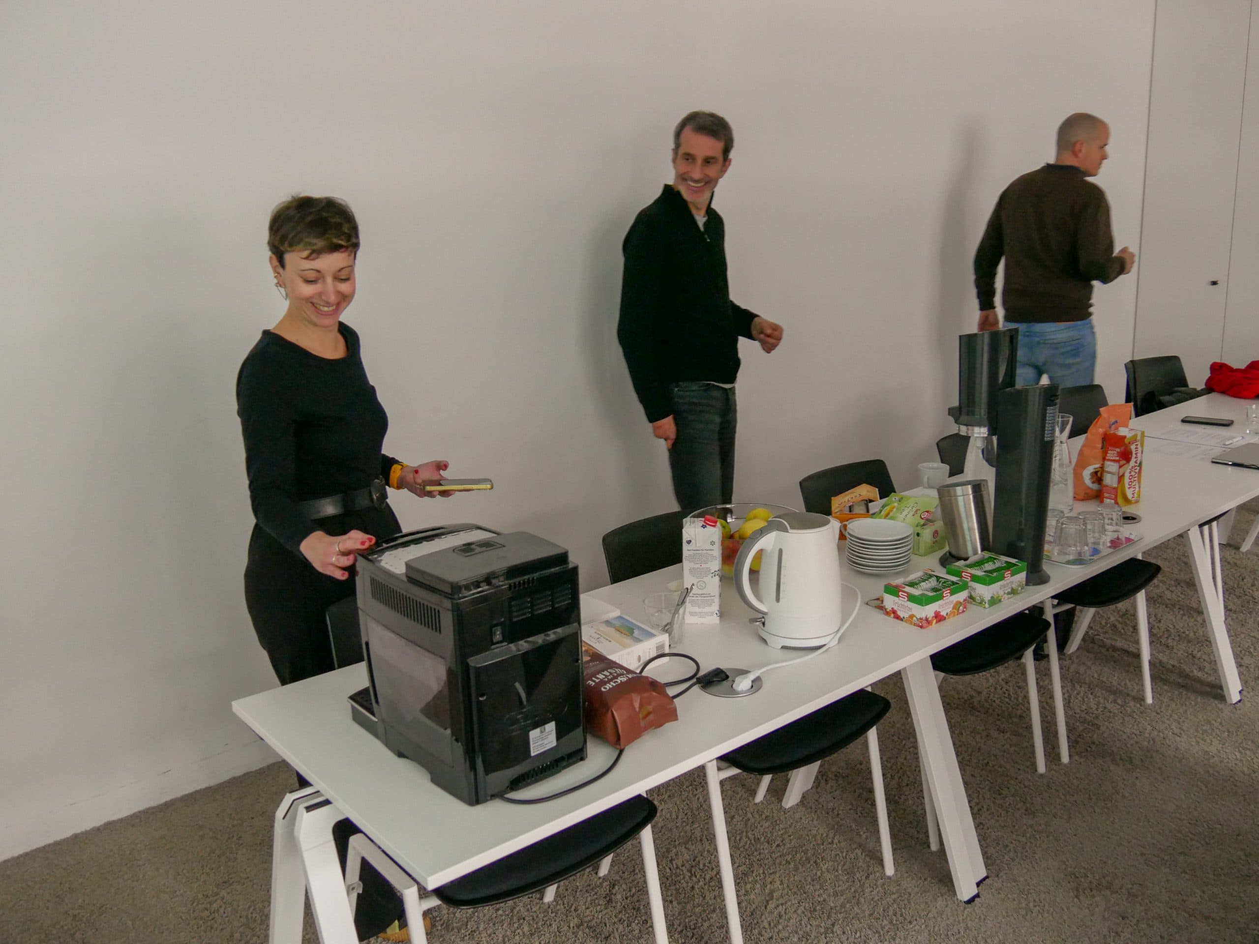 Drei Personen stehen an einem langen Tisch mit Kaffeemaschinen, Tassen, Snacks und Getränken. Eine Person bedient eine Kaffeemaschine, während die anderen lächeln und etwas zubereiten. Die Umgebung scheint ein zwangloses Büro oder ein Besprechungsraum zu sein.