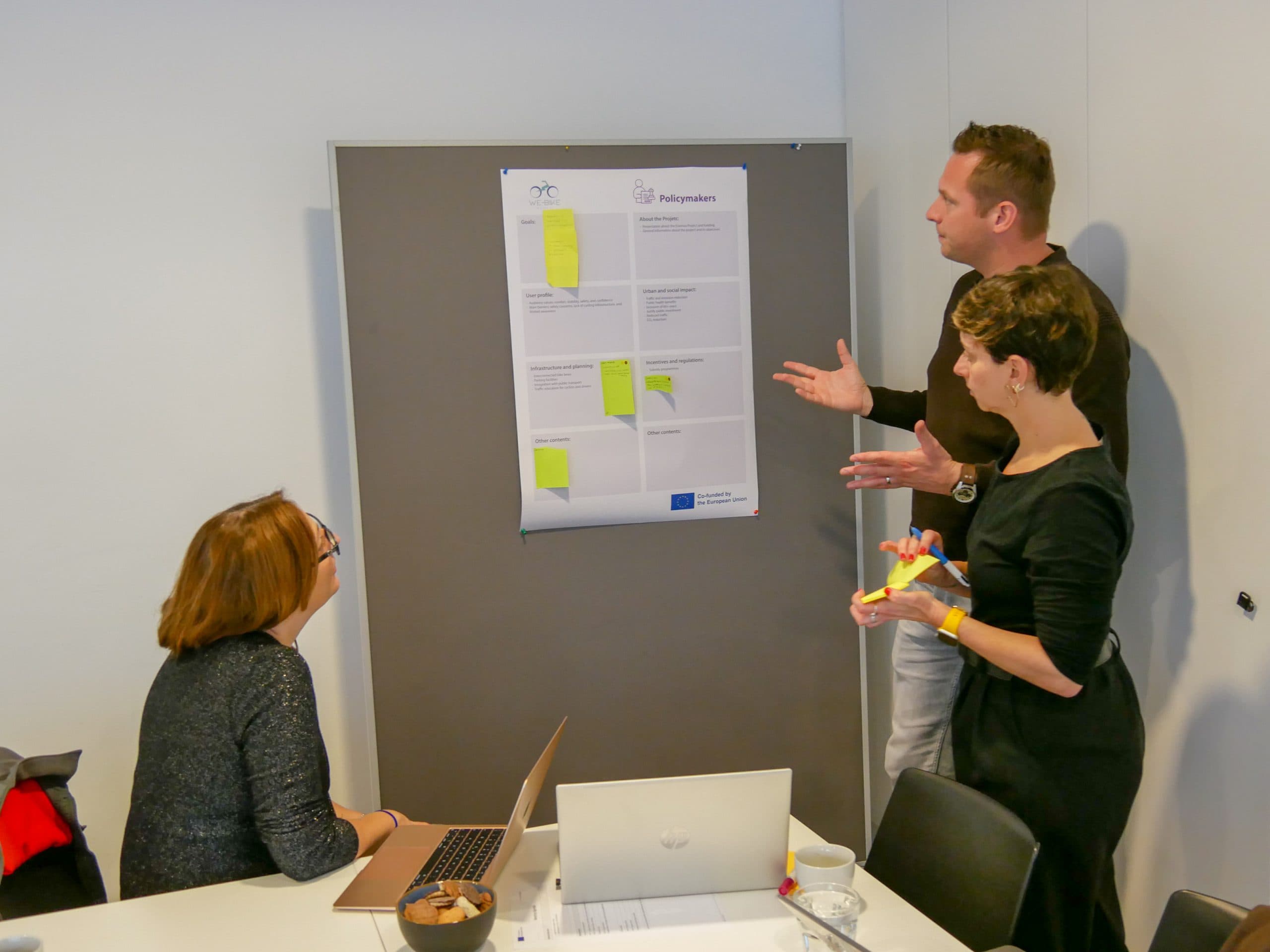 Drei Personen sitzen in einem Büro und diskutieren über ein großes Blatt mit Notizen an einer Tafel. Einer sitzt mit einem Laptop, während die beiden anderen stehen, gestikulieren und Stifte halten. Es scheint sich um eine gemeinsame Sitzung oder einen Workshop zu handeln.