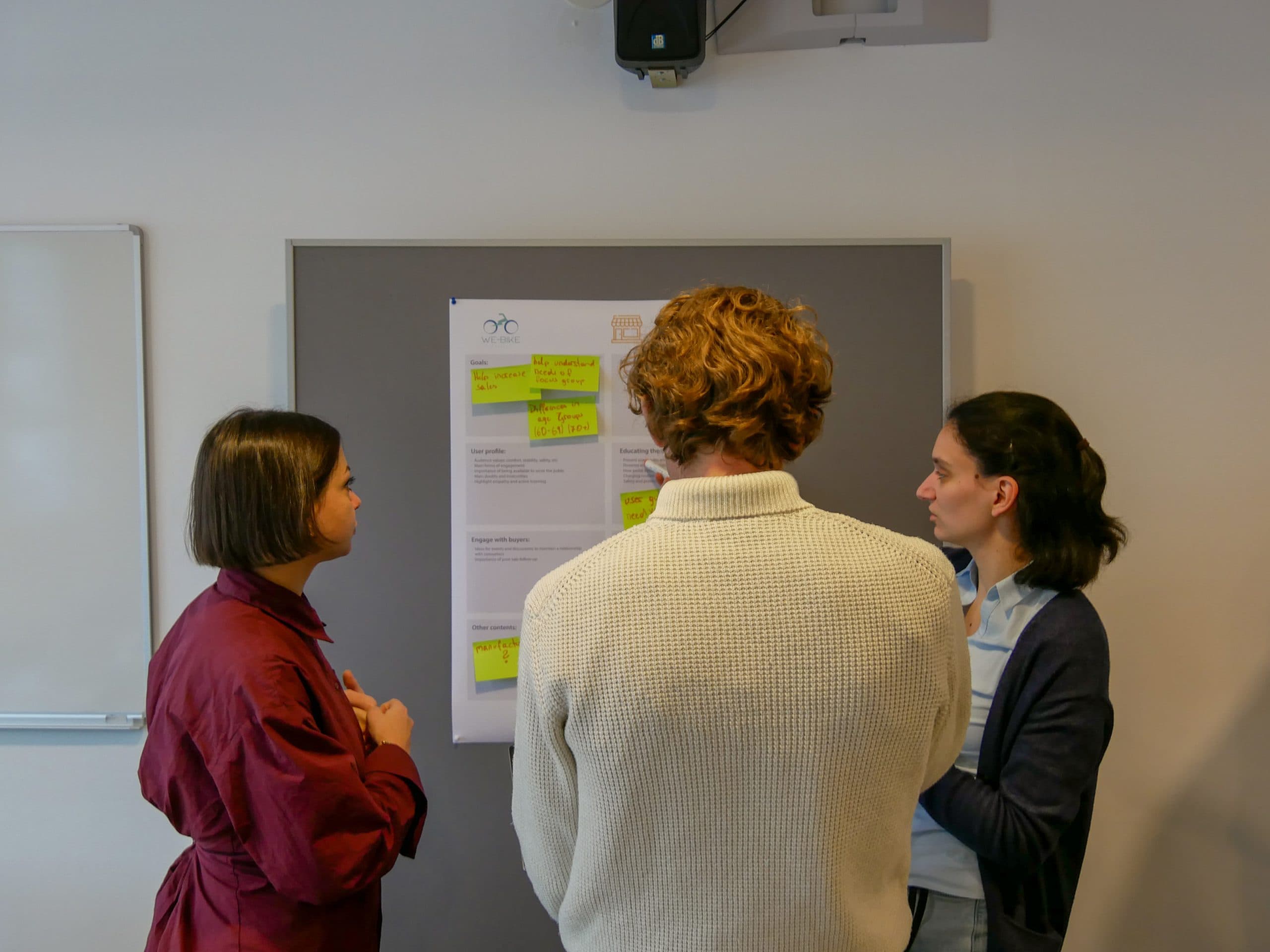 Drei Personen stehen in einem gut beleuchteten Raum vor einer Tafel mit Papier und Haftnotizen und diskutieren und arbeiten zusammen. Zwei von ihnen sind der Tafel zugewandt, während eine Person ihnen gegenübersteht und ein Gespräch führt.