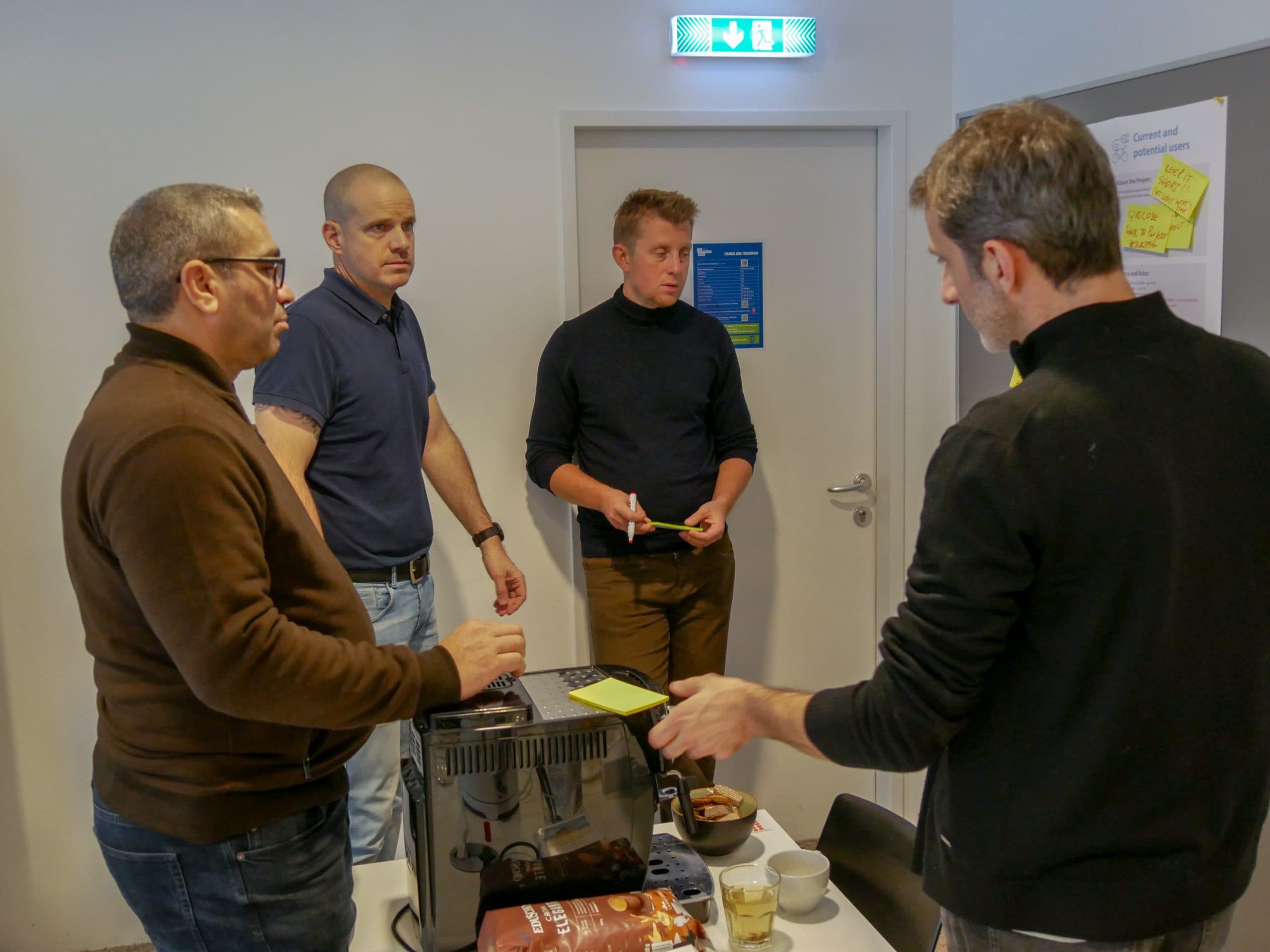 Vier Männer stehen um einen kleinen Tisch mit Snacks, Getränken und einer Kaffeemaschine und unterhalten sich in einem hell erleuchteten Raum mit Postern und Notizen an der Wand.