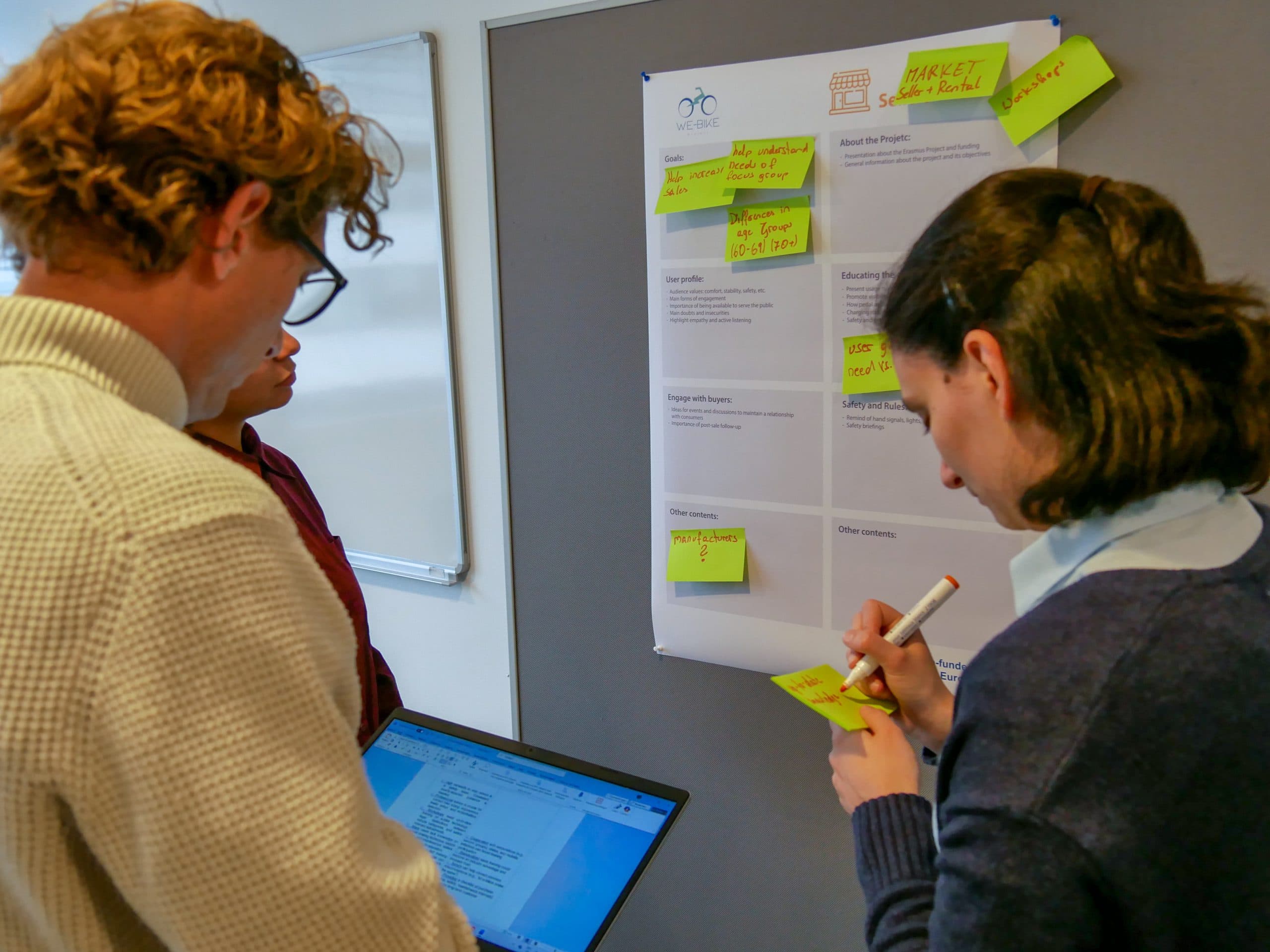 Drei Personen machen ein Brainstorming vor einer Tafel mit Haftnotizen. Einer schreibt auf einen Zettel, ein anderer hält einen Laptop, auf dem Text angezeigt wird, und die Projektdetails sind auf der Tafel sichtbar.