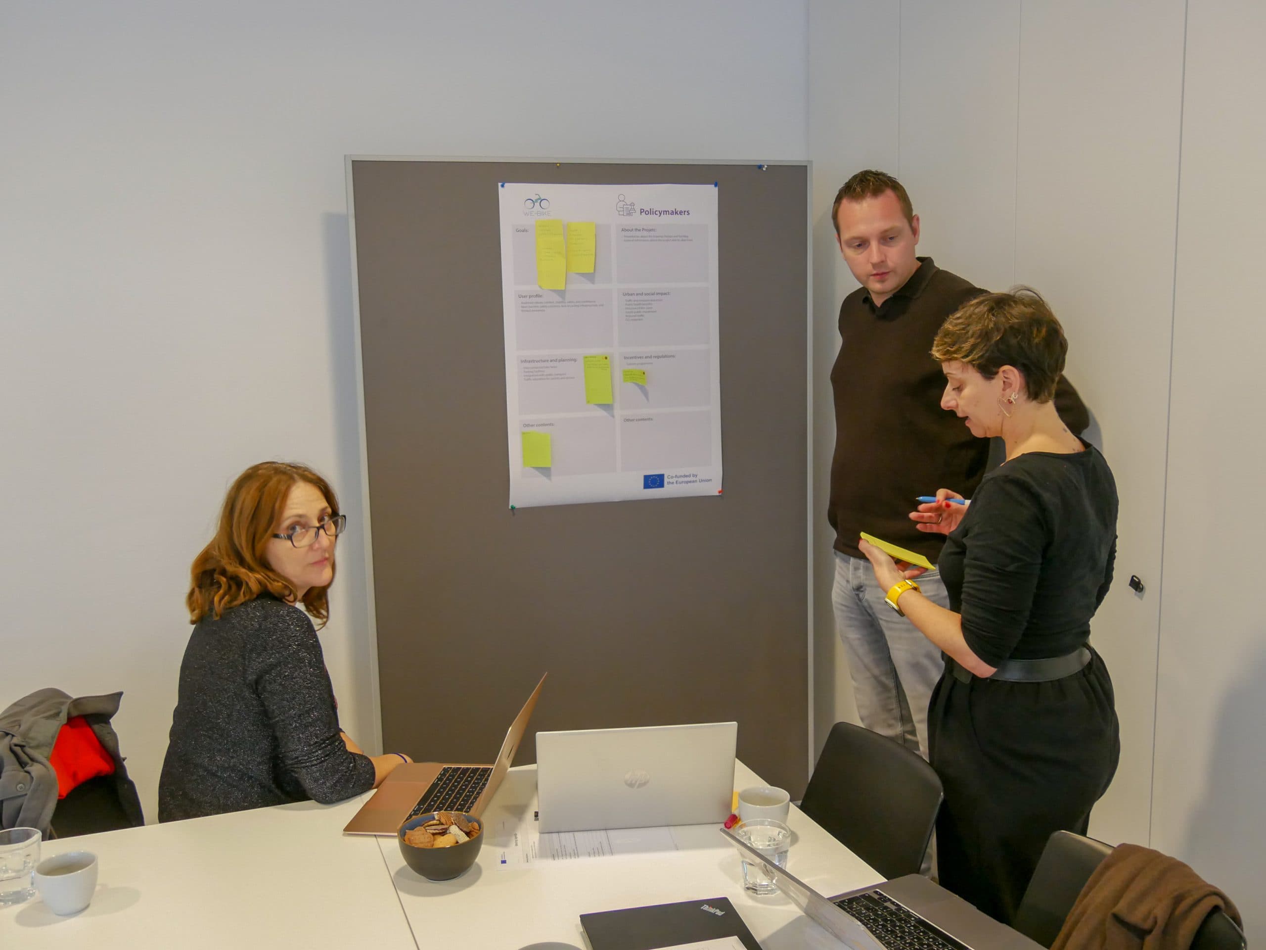 Drei Personen diskutieren in einem Büro über eine Tabelle mit Haftnotizen, die an einer Tafel angebracht sind. Zwei stehen und sprechen, während einer an einem Tisch mit Laptops und Kaffeetassen sitzt und in die Kamera schaut.