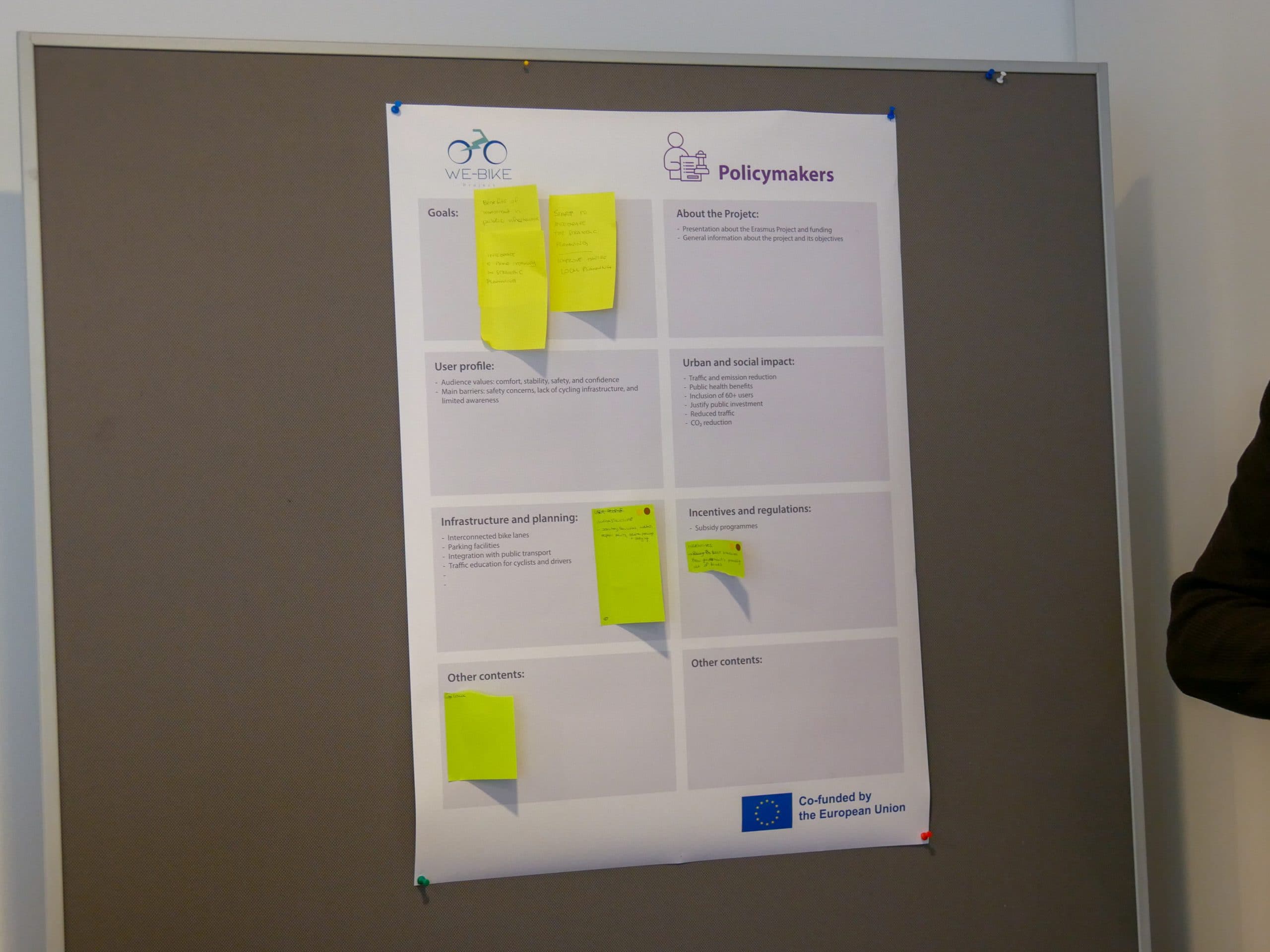 Ein Poster mit dem Titel 'Politische Entscheidungsträger' ist an eine graue Tafel gepinnt, auf der Abschnitte mit handgeschriebenen gelben Haftnotizen zu sehen sind. Das Poster wird von der Europäischen Union kofinanziert und informiert über Projektziele, Auswirkungen und Planung.