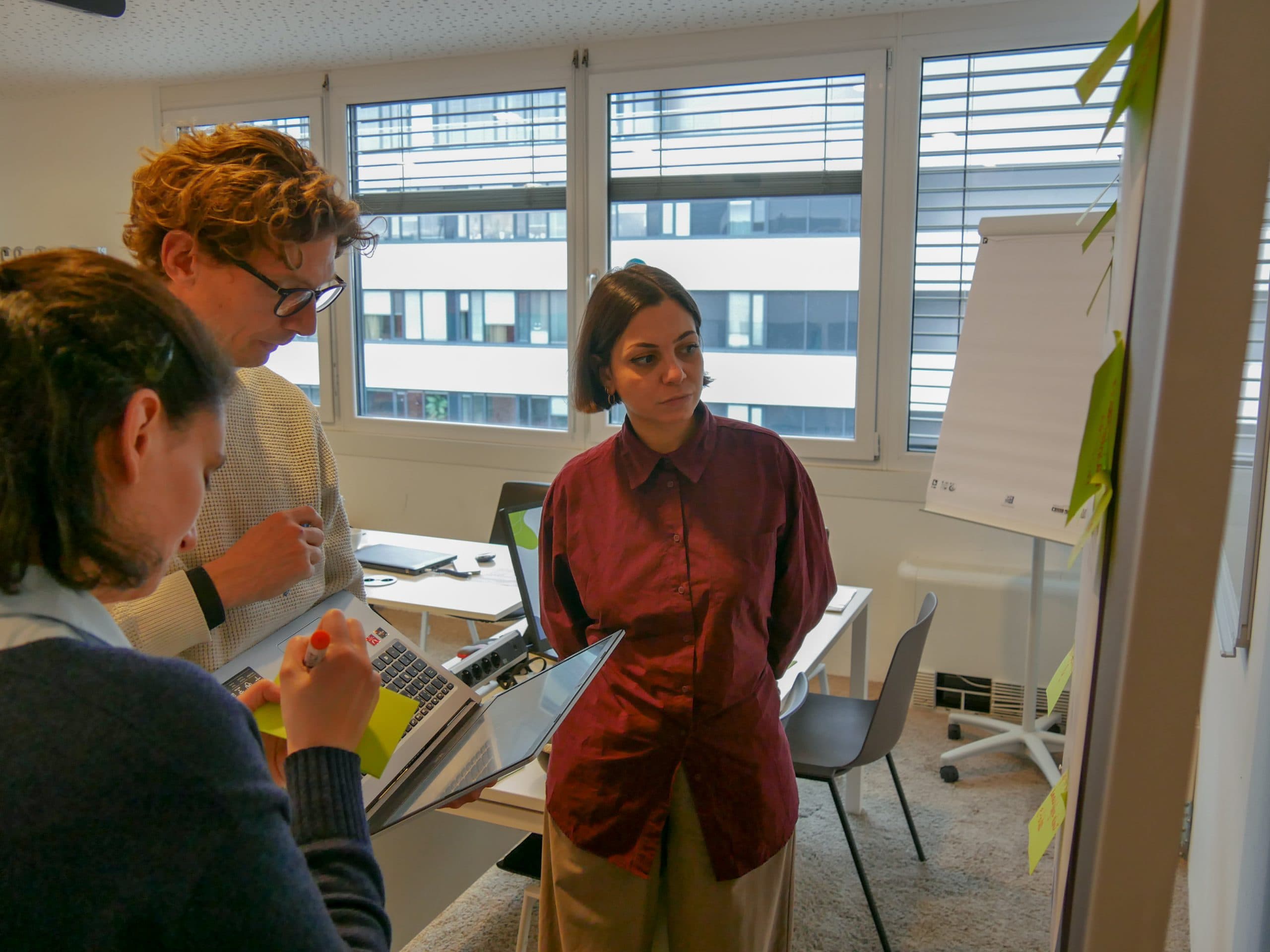 Drei Personen stehen in einem Büro und schauen auf Haftnotizen an der Wand. Eine Person hält einen Laptop, eine andere hält Haftnotizen und die dritte steht mit den Händen hinter dem Rücken. Im Hintergrund sind Tische und ein Flipchart zu sehen.