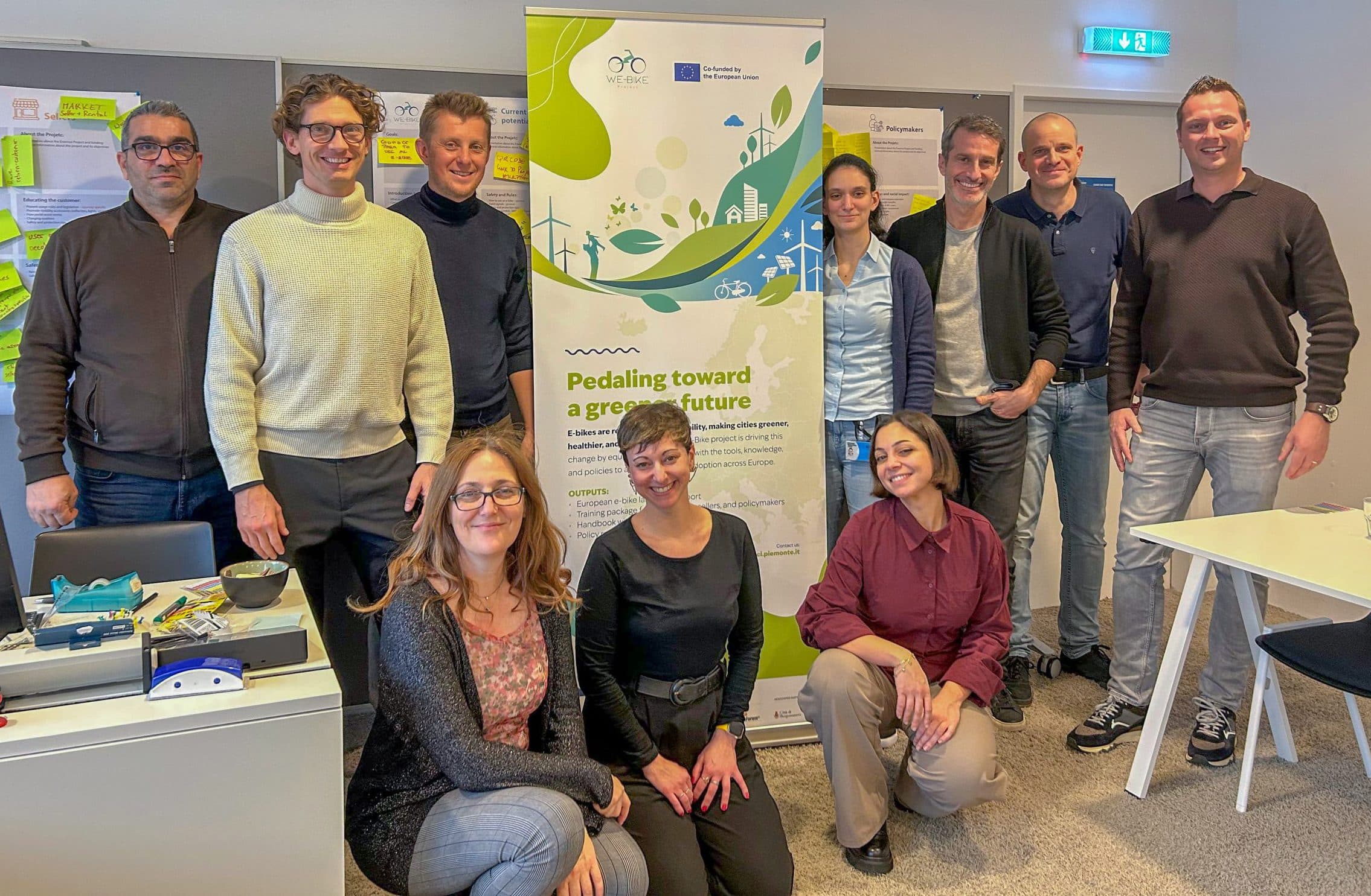 Eine Gruppe von zehn Personen posiert und lächelt in einem Büro, wobei einige stehen und andere knien. Hinter ihnen hängt ein Banner mit der Aufschrift "Pedalling towards a greener future", und an der Wand sind bunte Klebezettel angebracht.