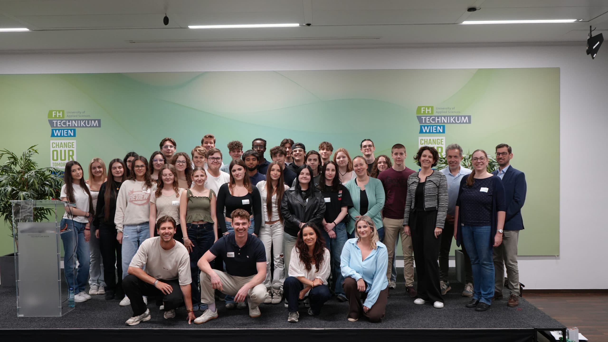 Eine große Gruppe von Menschen, meist junge Erwachsene, posiert und lächelt gemeinsam vor einem grün-blauen Hintergrund mit der Aufschrift FH Technikum Wien und Change Our World bei einer Indoor-Veranstaltung.