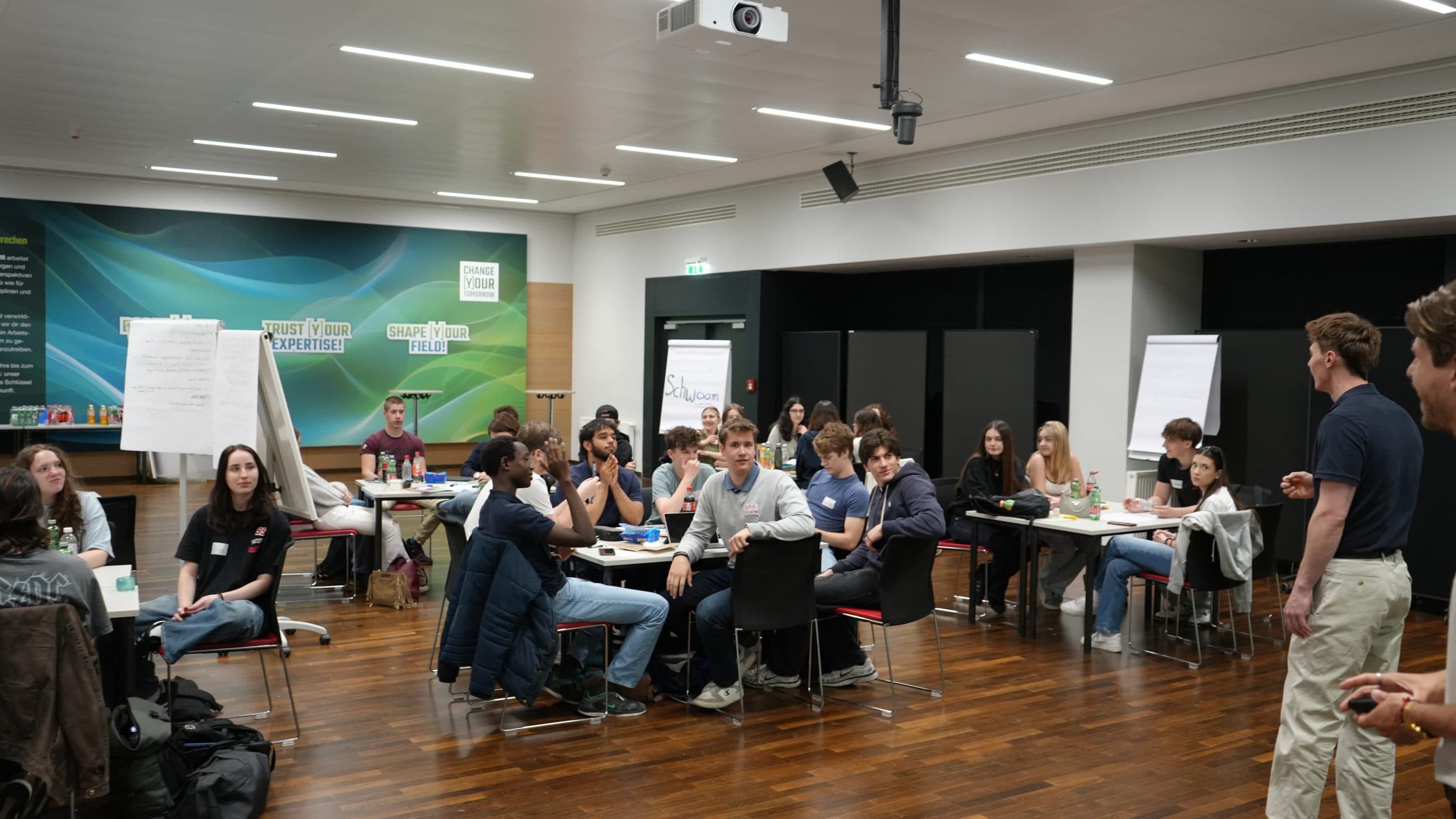 Eine Gruppe junger Erwachsener sitzt an Tischen in einem modernen Tagungsraum und hört einem Redner zu. Flipcharts und Material sind sichtbar, und ein Projektor hängt von der Decke. Die Atmosphäre wirkt kooperativ und konzentriert.