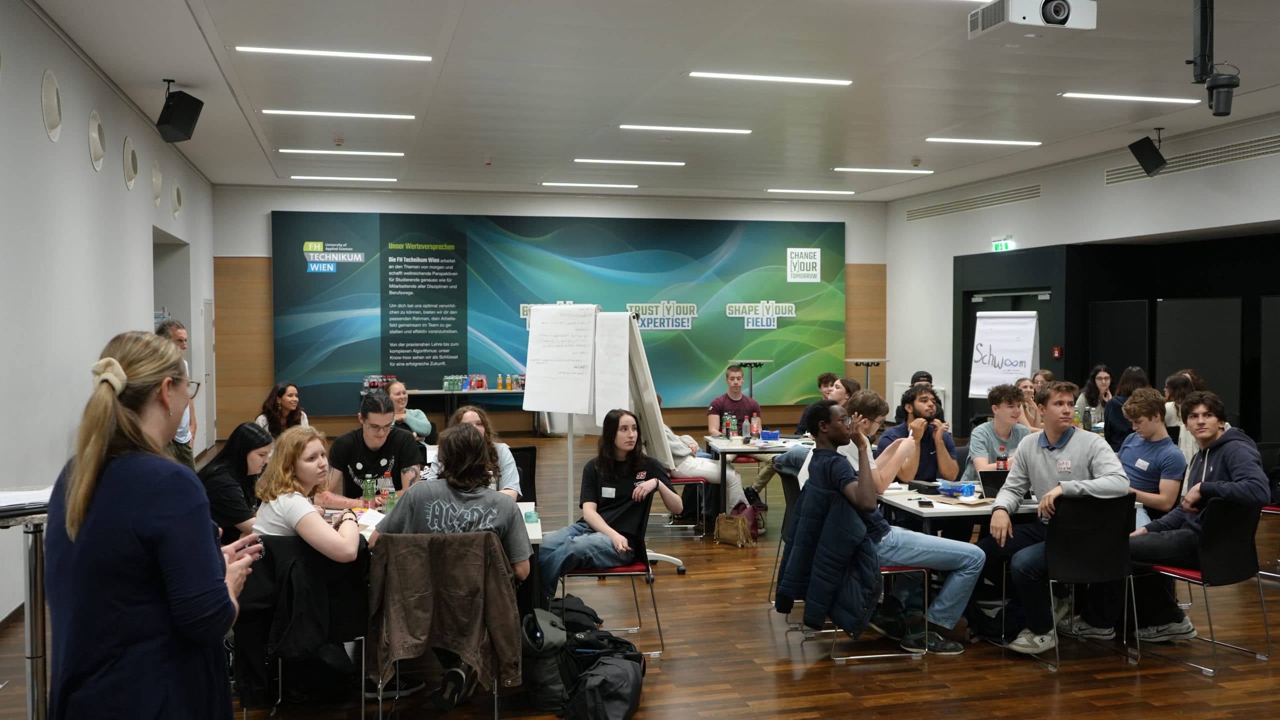 Eine Gruppe von Personen unterschiedlichen Alters sitzt an Tischen in einem modernen Konferenzraum und hört einem stehenden Redner zu. Im Hintergrund sind ein großes Plakat und ein Flipchart zu sehen.