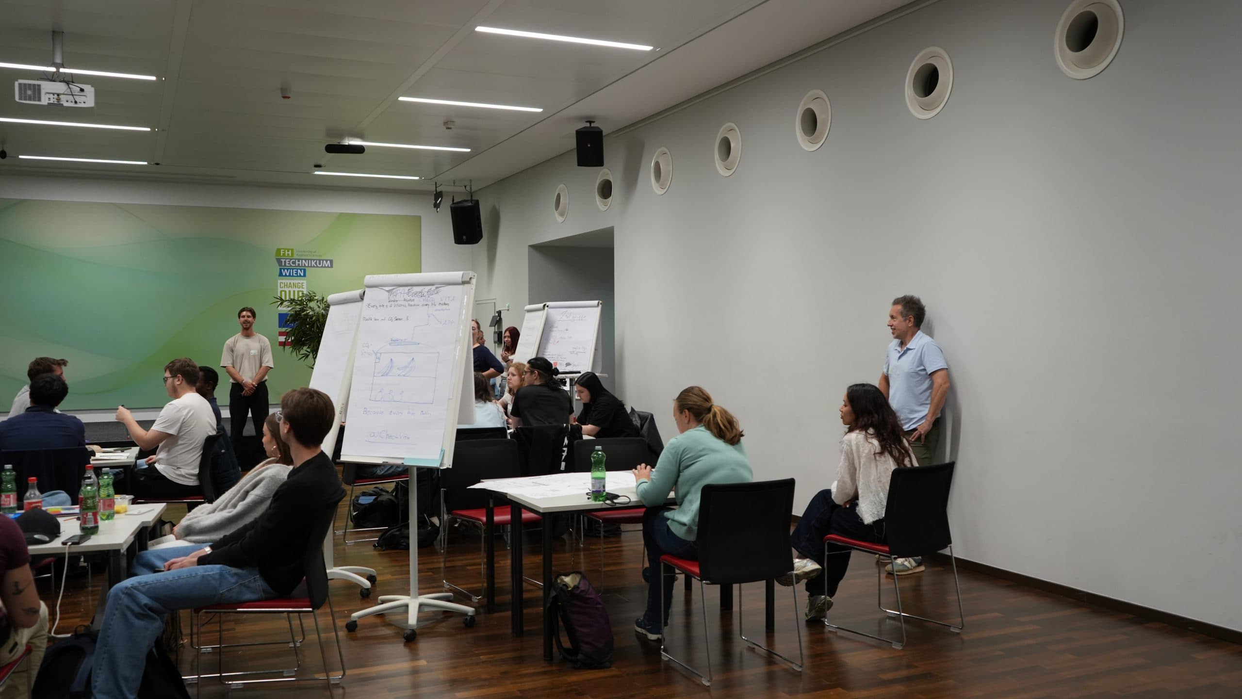 Eine Gruppe von Personen sitzt in einem modernen Klassenzimmer an Tischen und vor Flipcharts mit Diagrammen. Einige Personen stehen in der Nähe der Tafeln, während andere aufmerksam zuhören. Der Raum hat weiße Wände und Holzböden.
