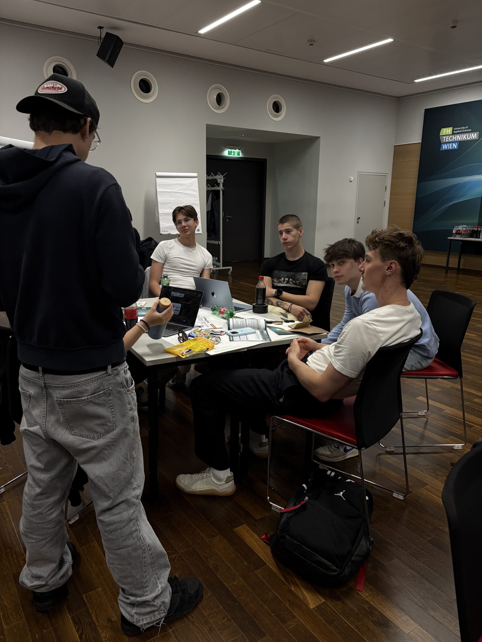 Fünf junge Männer sitzen in einem modernen Raum an einem Tisch, auf dem Laptops und Getränke verteilt sind.