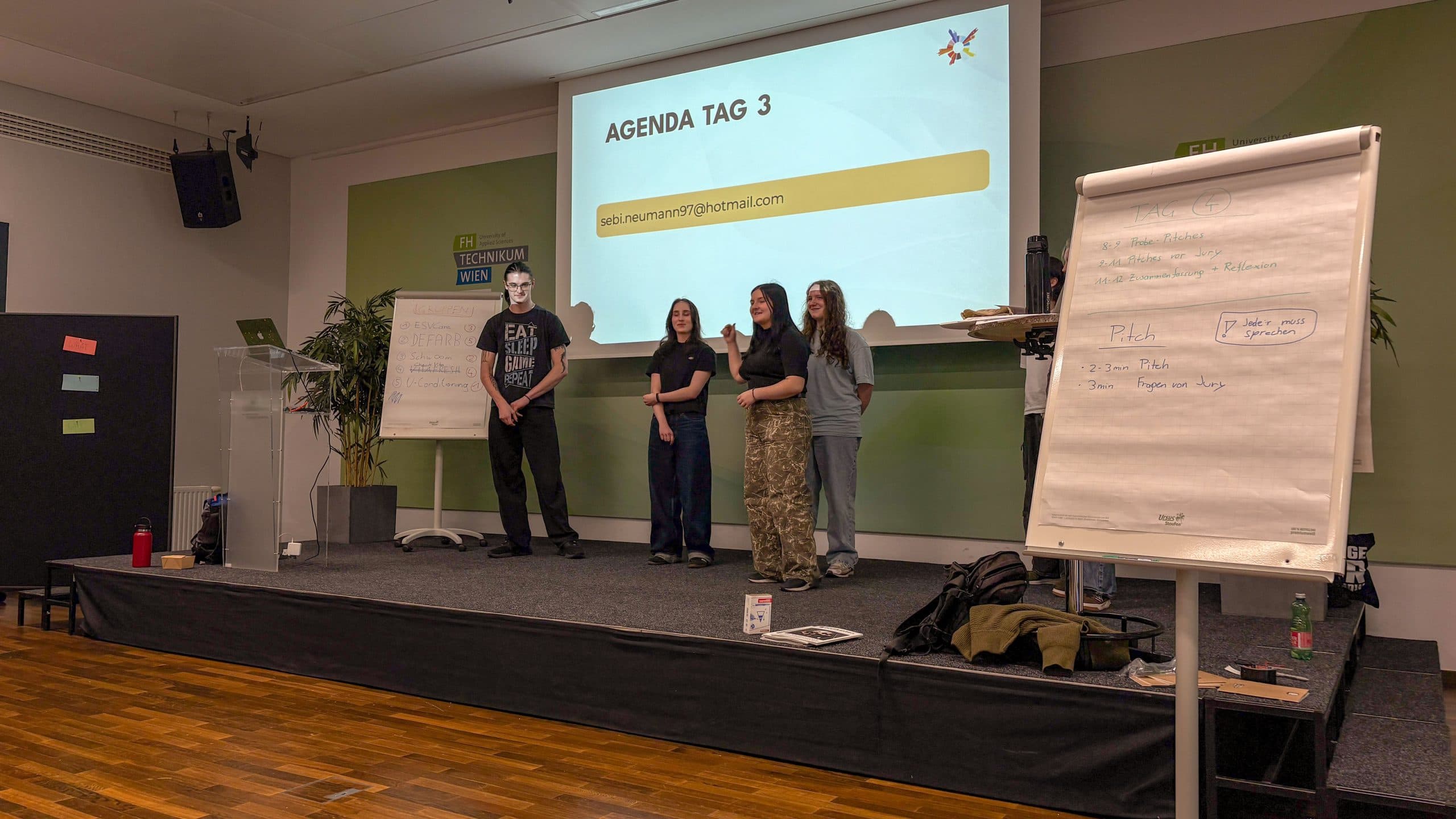 Menschen auf der Bühne vor einem Projektionsschirm mit dem Text AGENDA TAG 3 und einer Tafel mit TAG 4 darauf.