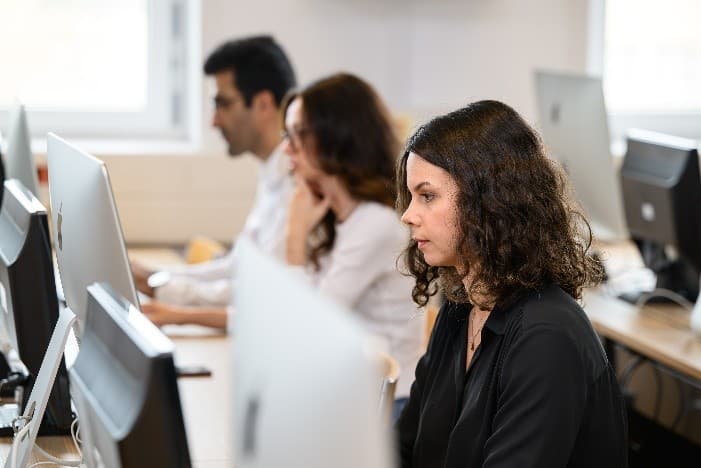 Drei Personen sitzen an Schreibtischen und arbeiten an Desktop-Computern in einem hellen Büro oder Klassenzimmer. Der Fokus liegt auf einer Frau mit lockigem Haar im Vordergrund; die anderen sind im Hintergrund leicht verschwommen.