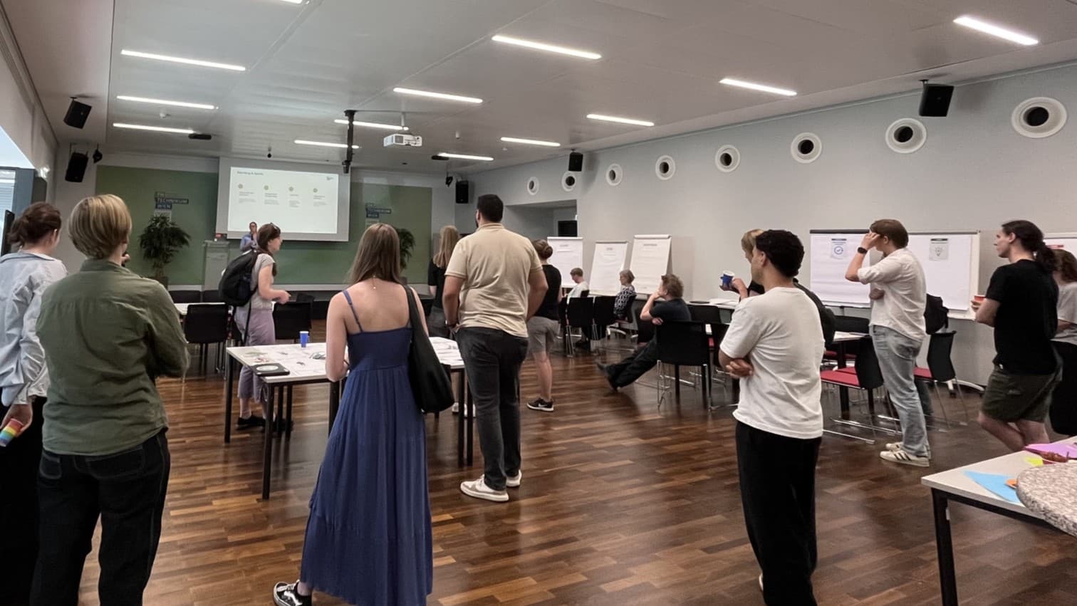Eine Gruppe von Personen steht und sitzt in einem modernen, hell erleuchteten Konferenzraum und verfolgt eine Präsentation, die auf eine Leinwand an der Stirnseite projiziert wird. Mehrere Poster und Flipcharts sind im Raum verteilt.