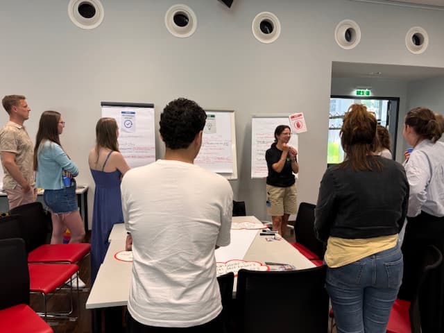 Eine Gruppe von Personen steht in einem modernen Sitzungssaal und hört einem Vortragenden zu, der drei Flipcharts mit Notizen vor sich hat. Einige machen sich Notizen, andere schauen aufmerksam zu. Die Umgebung wirkt kooperativ und informell.