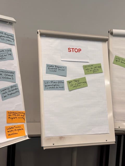Ein Flipchart mit einem STOP-Zeichen am oberen Rand und mehreren farbigen Notizen, auf denen verschiedene handschriftliche Aufgaben oder Themen aufgeführt sind. Auf der linken Seite ist ein weiteres Flipchart mit weiteren farbigen Notizen zu sehen.