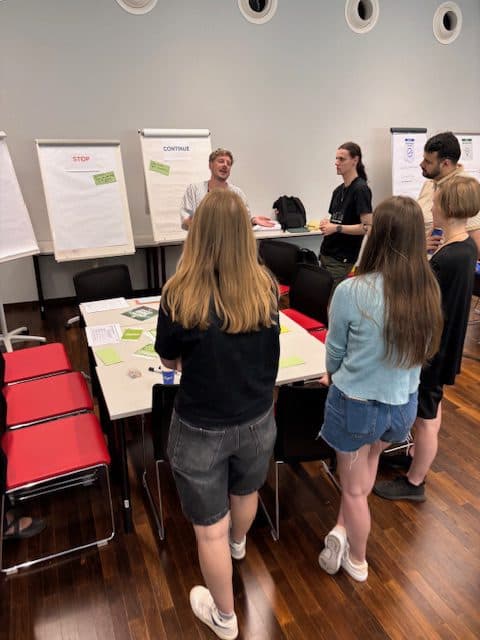 Eine Gruppe von fünf Personen steht in einem Besprechungsraum um einen Tisch herum und hört einem Redner zu. Hinter ihnen sind Flipcharts mit Notizen aufgestellt, und auf dem Tisch liegen Papiere aus.
