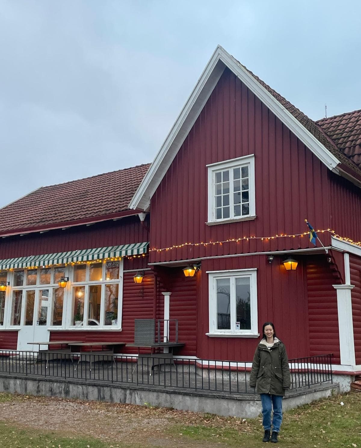 Eine Person in einem grünen Mantel steht vor einem großen roten Holzhaus mit weißer Verkleidung und einem Ziegeldach. Das Haus hat Außenbeleuchtung, eine Fahne und eine Bank, im Hintergrund ist der Himmel bewölkt.