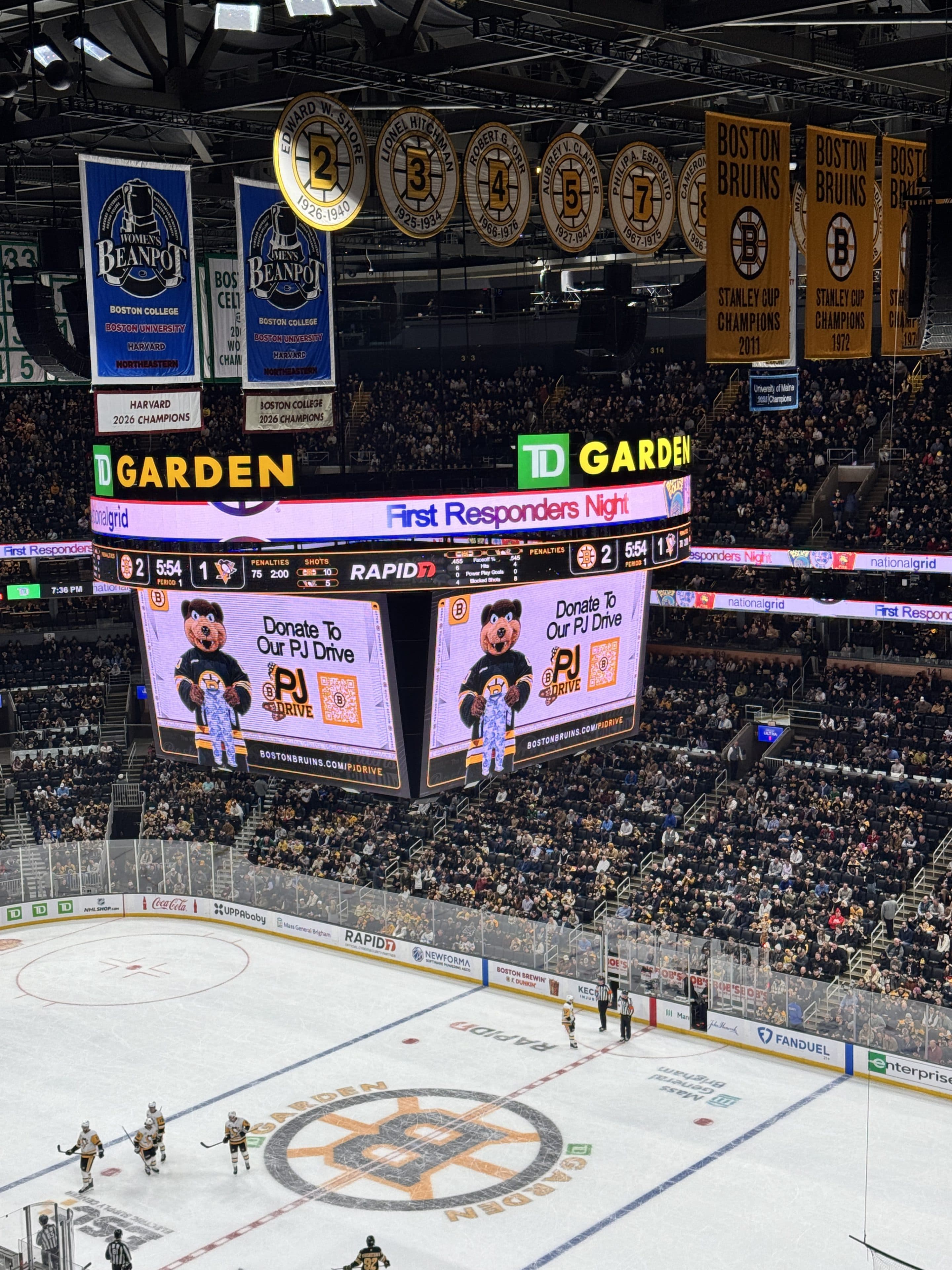 Ein volles Haus im TD Garden verfolgt ein Eishockeyspiel. Auf der Anzeigetafel steht "Donate to Our PJ Drive" mit einem Cartoon-Bären, und von den Dachsparren hängen Banner für die Boston Bruins.