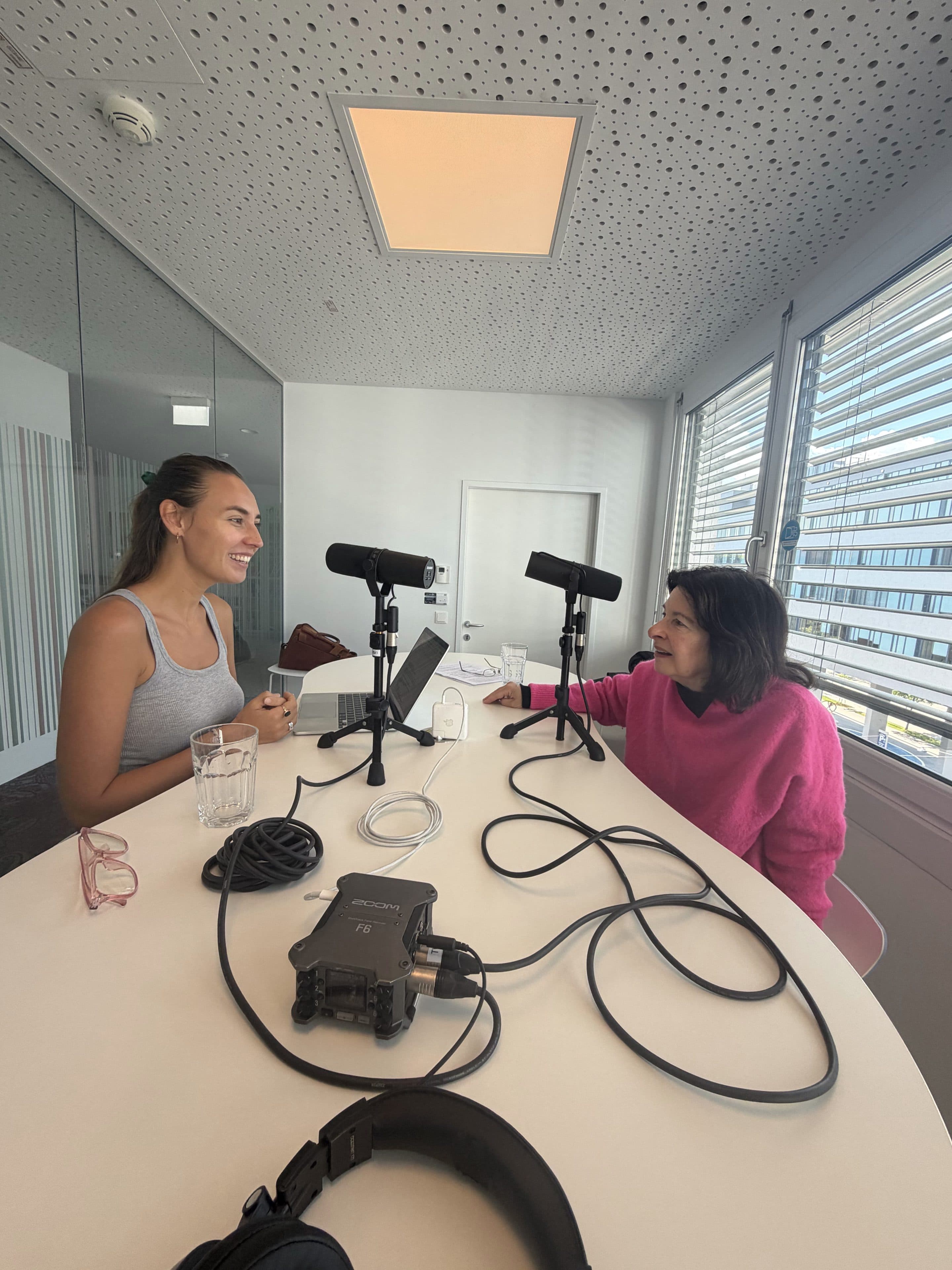 In einem modernen, hellen Büro mit großen Fenstern und einer Glaswand sitzen zwei Frauen an einem Tisch mit Mikrofonen und Aufnahmegeräten zwischen ihnen, lächeln und reden.