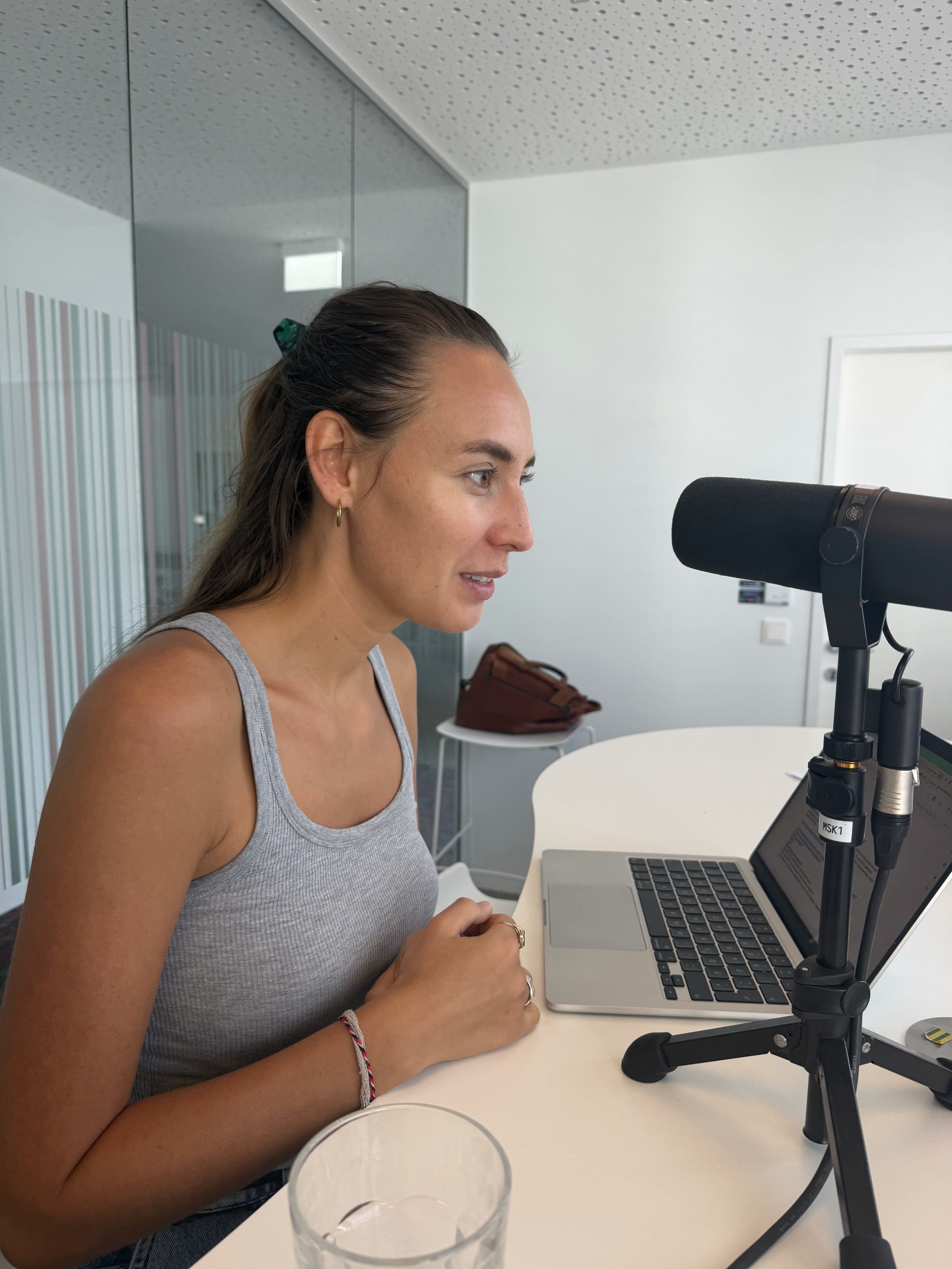Eine Frau in einem grauen Tank-Top spricht in ein Mikrofon, während sie an einem Schreibtisch mit einem Laptop sitzt. Ein Glas Wasser und eine braune Tasche stehen auf dem Schreibtisch. Es scheint, als würde sie in einem modernen Büro aufnehmen oder streamen.