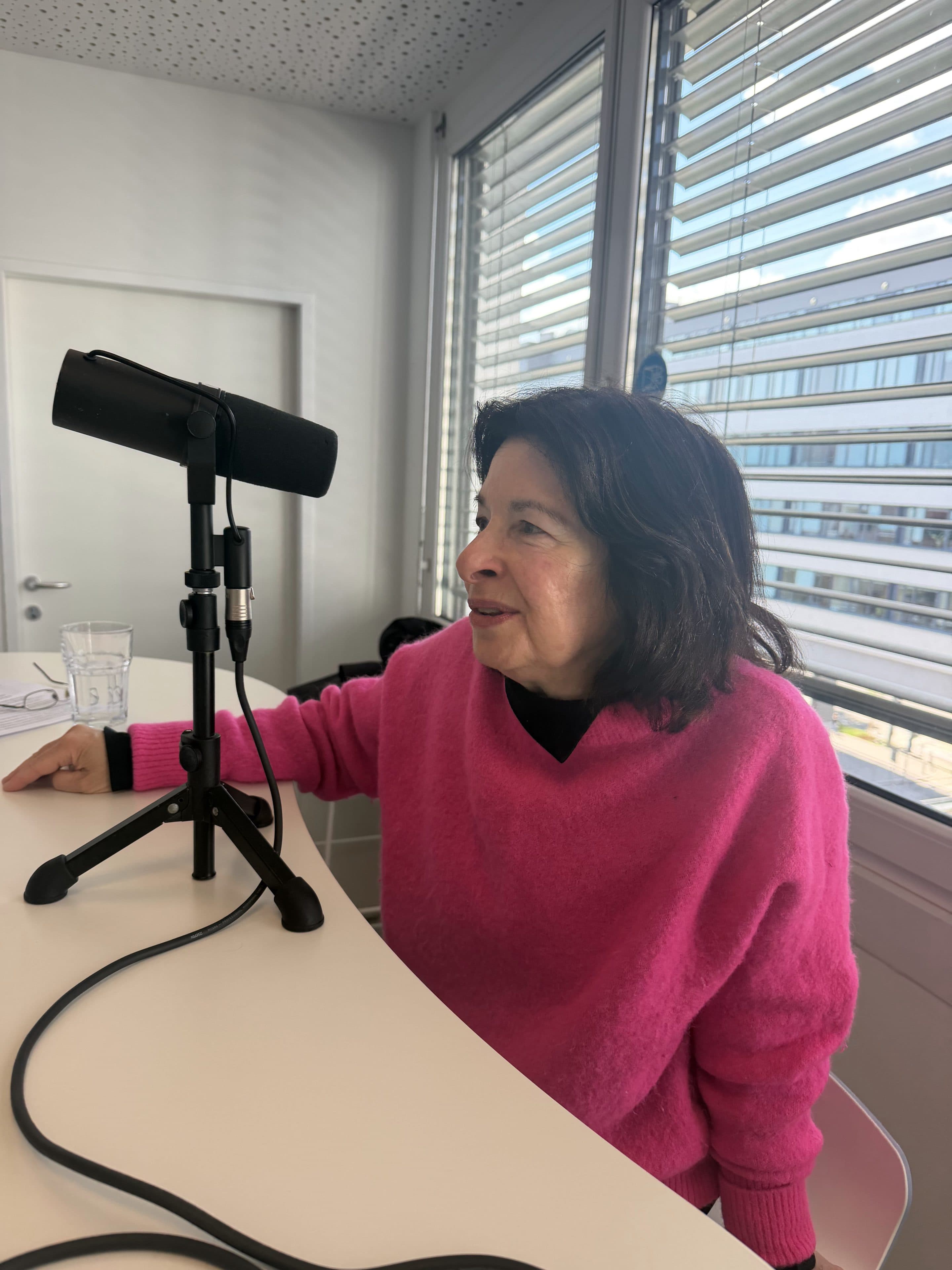 Eine Frau in einem hellrosa Pullover spricht in ein Mikrofon auf einem Tisch in einem modernen Büro mit großen Fenstern und Jalousien.