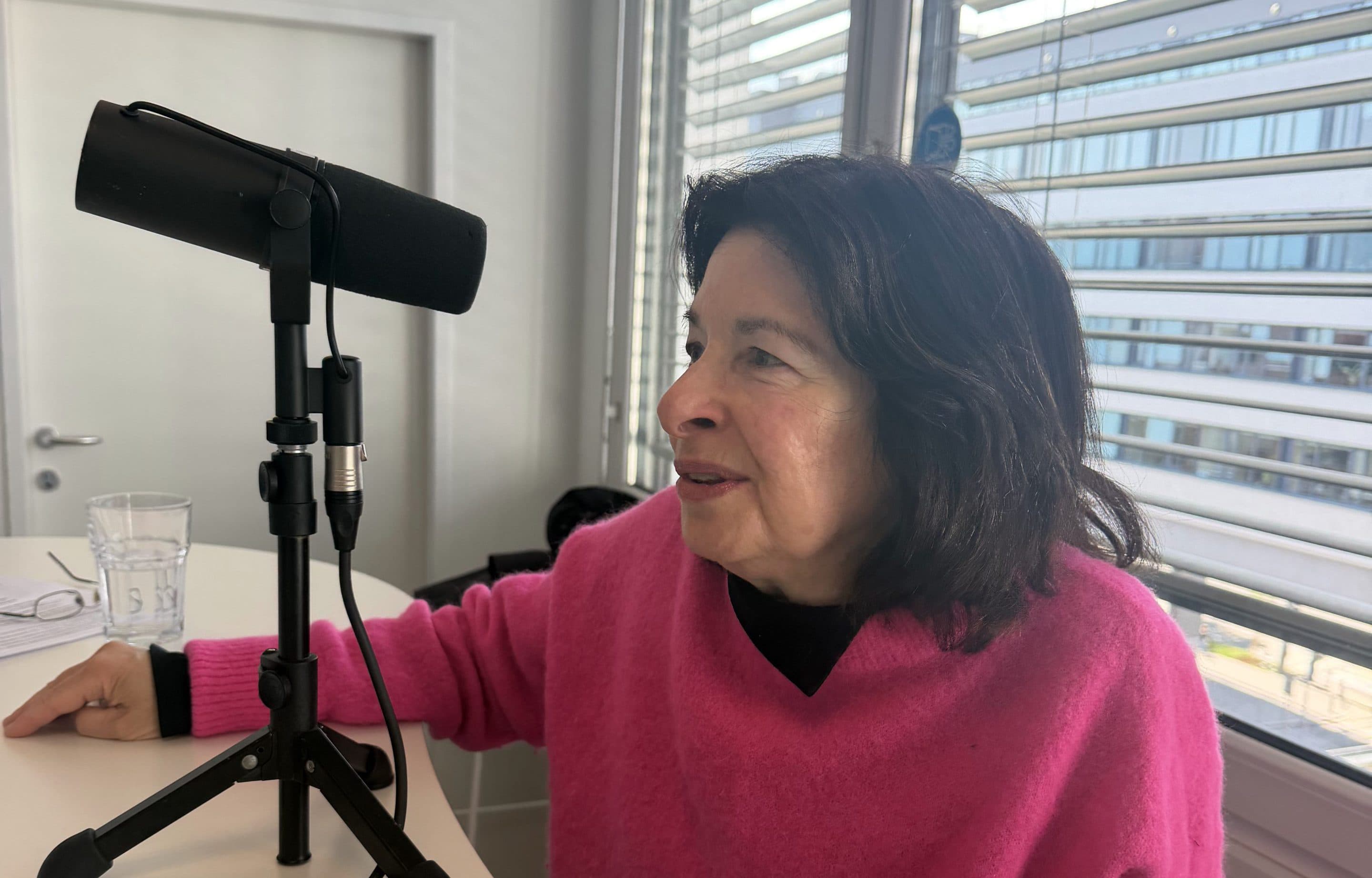 Eine Frau in einem hellrosa Pullover sitzt an einem Schreibtisch und spricht in ein Mikrofon. Sie befindet sich in einem modernen, sonnendurchfluteten Raum mit großen Fenstern und horizontalen Jalousien. Ein Glas Wasser und Kabel stehen auf dem Schreibtisch.