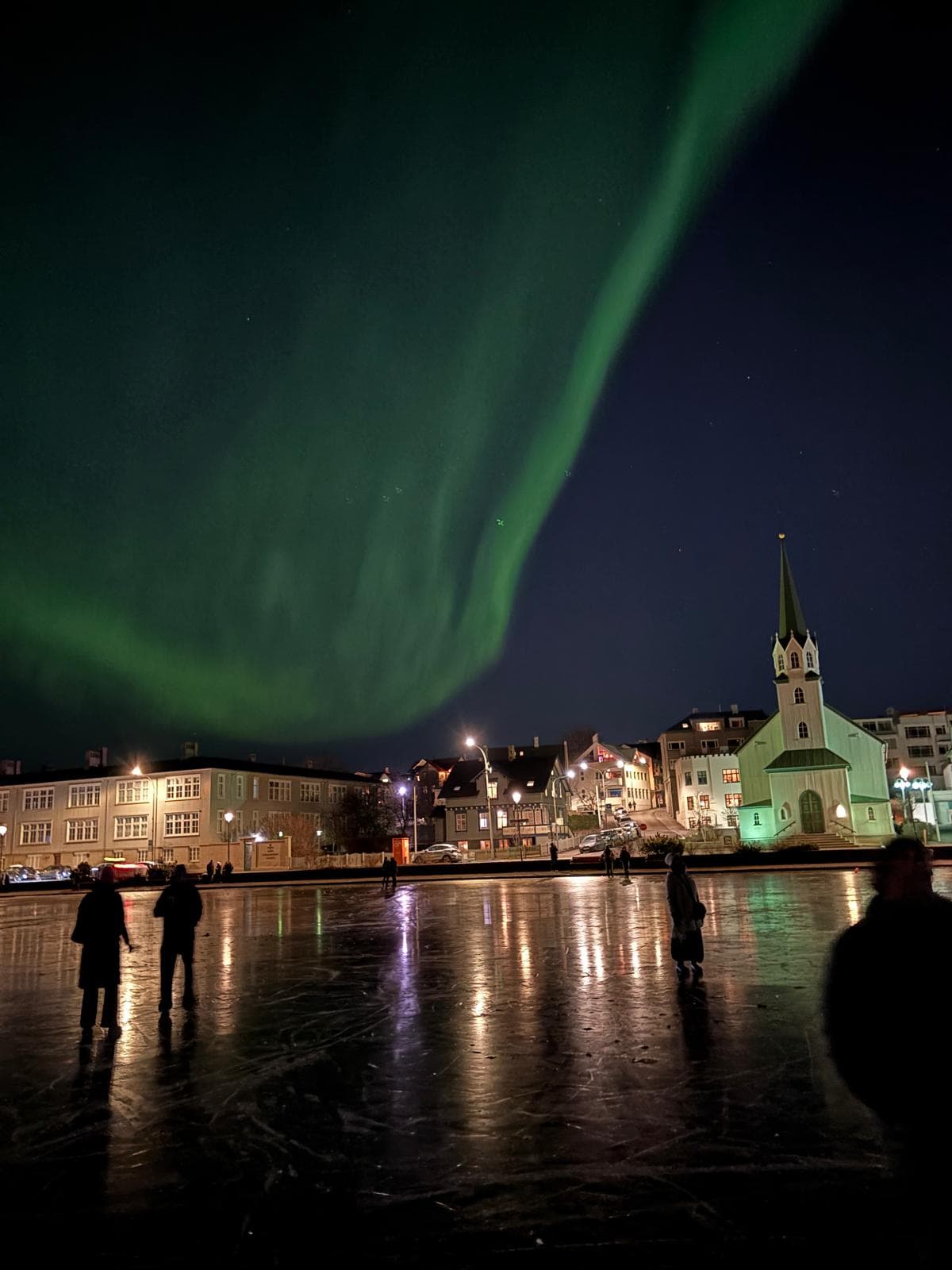 Menschen stehen nachts auf einer vereisten Fläche, die sich von Gebäuden und einer Kirche abhebt. Über ihnen erstrecken sich leuchtend grüne Nordlichter über den dunklen Himmel.