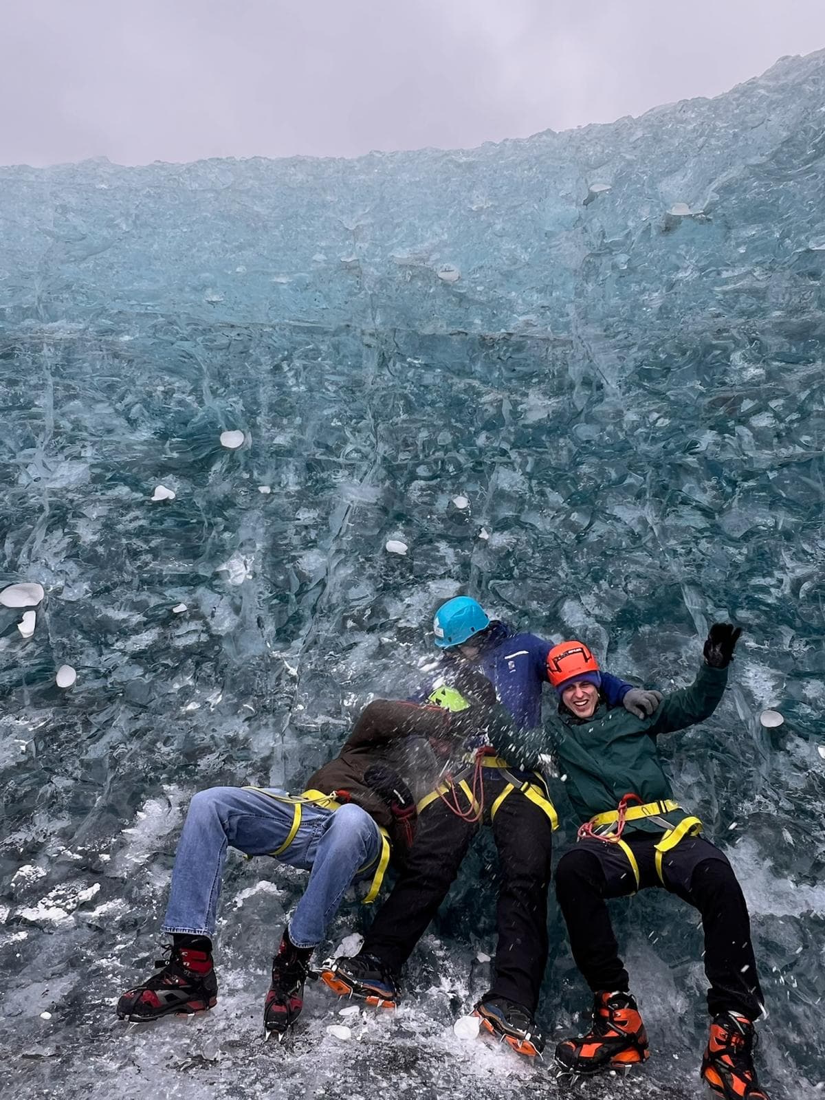 Drei Personen mit Helmen und Kletterausrüstung sitzen vor einer massiven Wand aus blauem Eis, lächeln und posieren für die Kamera. Die Gruppe scheint ein Outdoor-Abenteuer in einer gefrorenen, eisigen Umgebung zu genießen.