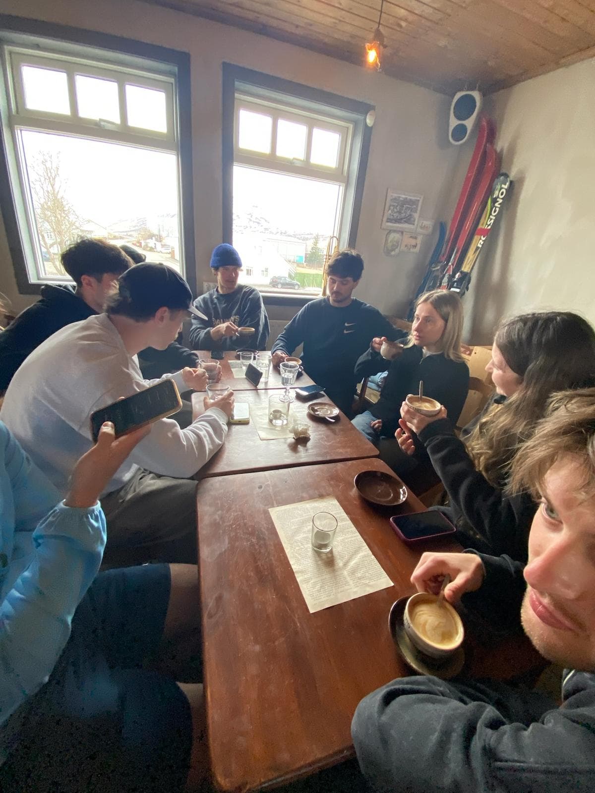 Eine Gruppe von acht jungen Erwachsenen sitzt um einen Holztisch in einem gemütlichen Café, trinkt Kaffee und unterhält sich. Große Fenster lassen das Tageslicht herein, und an der Wand hinter ihnen lehnt eine Skiausrüstung.