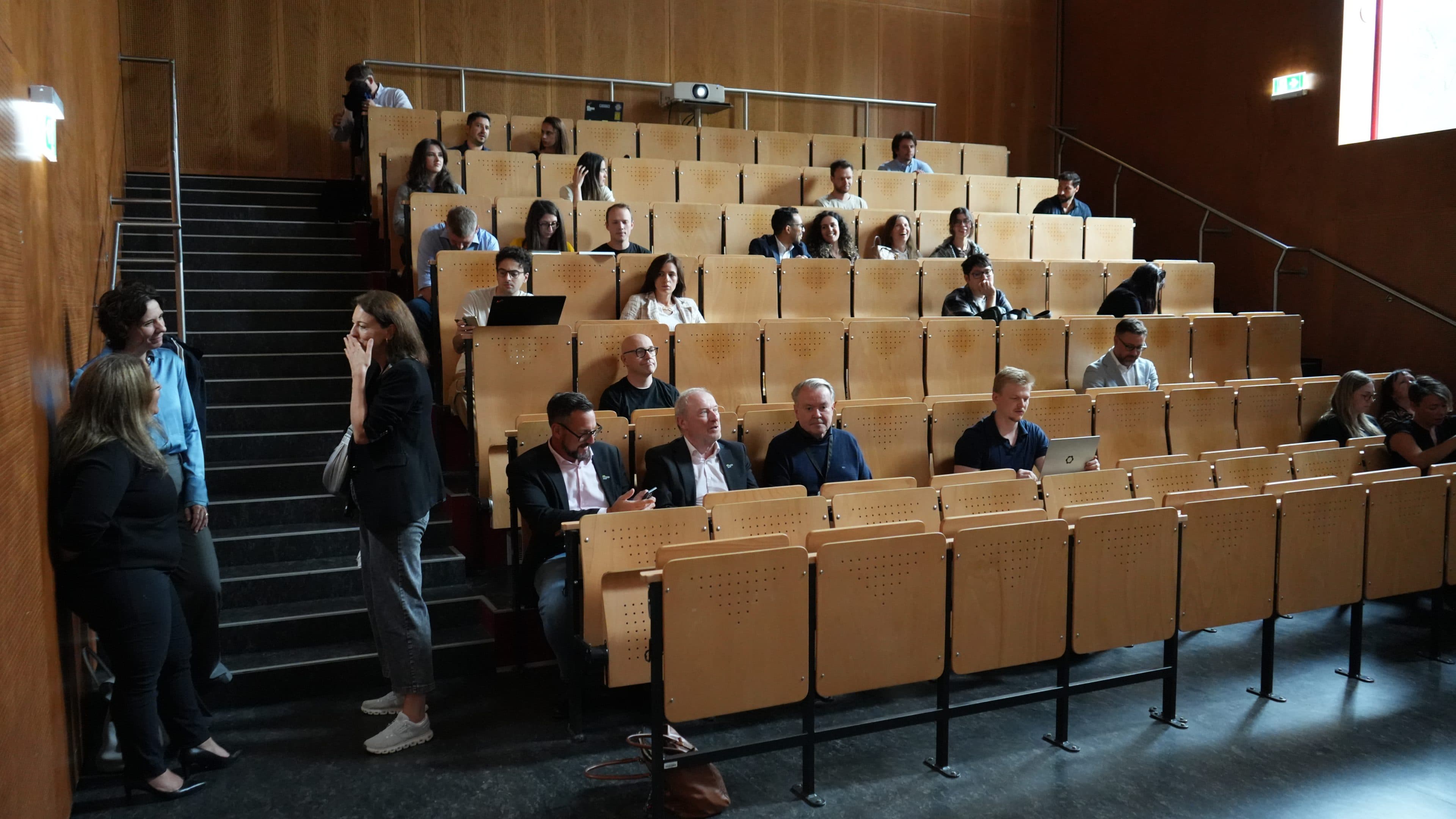 Eine Gruppe von Menschen sitzt verstreut auf Holzstühlen in einem Hörsaal; einige unterhalten sich, arbeiten am Laptop oder warten. Ein paar Personen stehen am Gang und unterhalten sich. Der Raum hat holzvertäfelte Wände und wird von Tageslicht durchflutet.