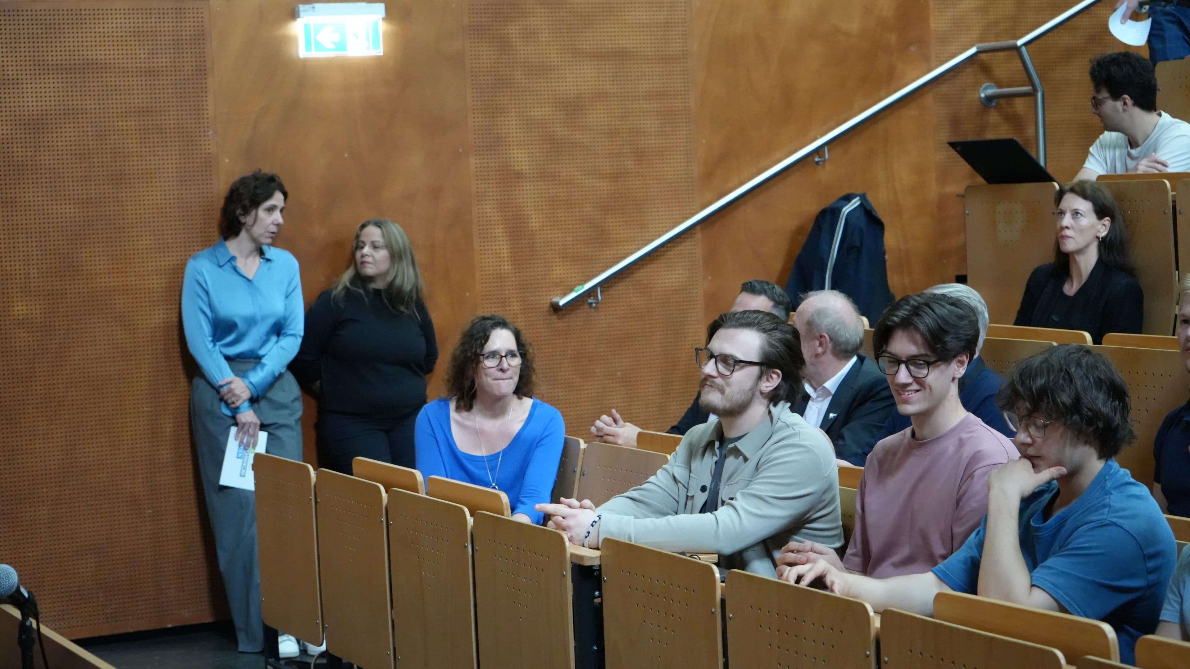 In einem Hörsaal mit Holzsitzen und -wänden sitzen mehrere Menschen und schauen aufmerksam zu; zwei Frauen stehen an der Seite, von denen eine Unterlagen in der Hand hält, während andere sitzen und entweder konzentriert oder entspannt wirken.
