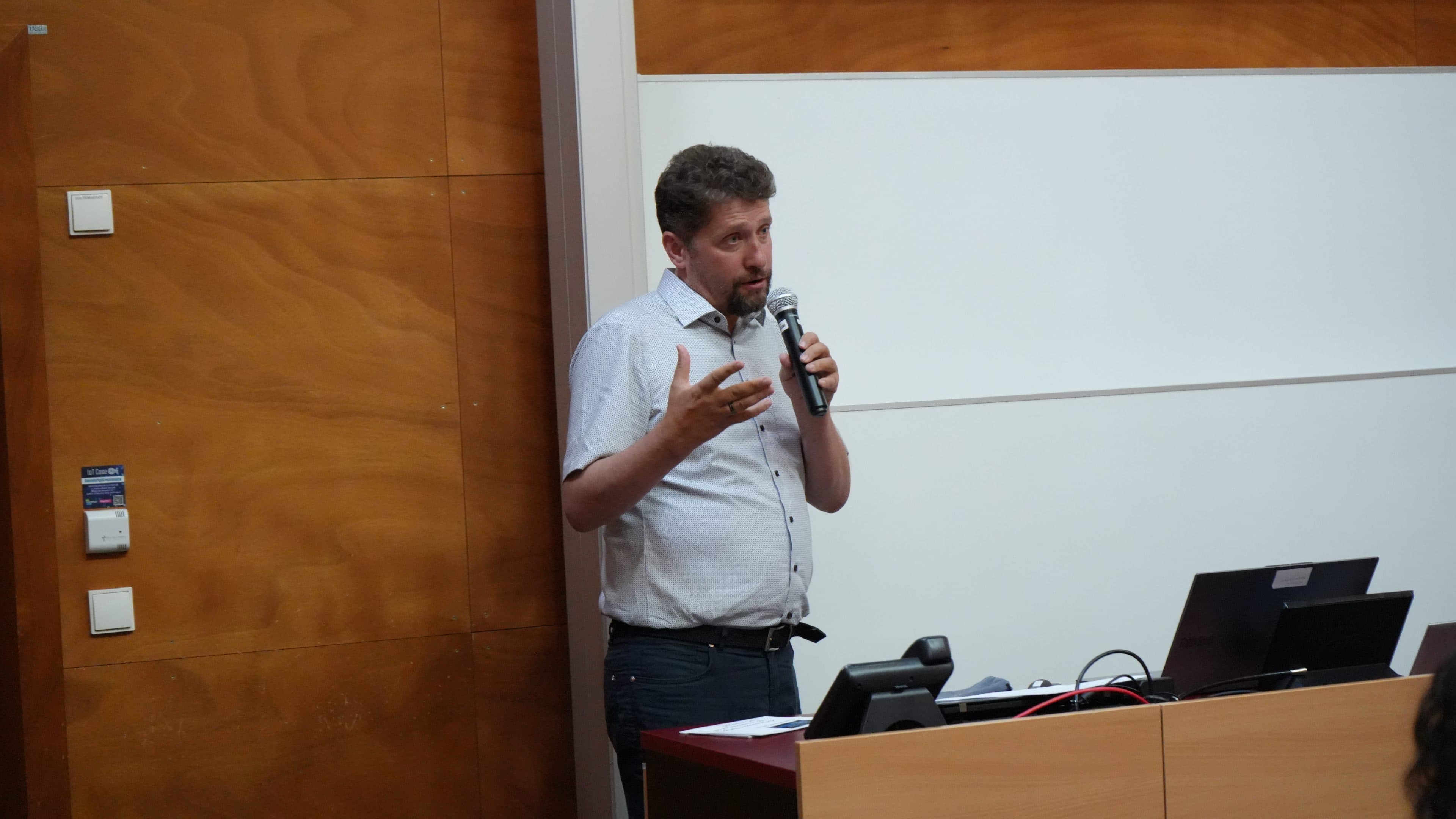 Ein Mann steht an einem Rednerpult, hält ein Mikrofon in der Hand und gestikuliert beim Sprechen. Er befindet sich in einem Raum mit holzgetäfelten und weißen Wänden; auf dem Rednerpult stehen ein Laptop und weitere Geräte.
