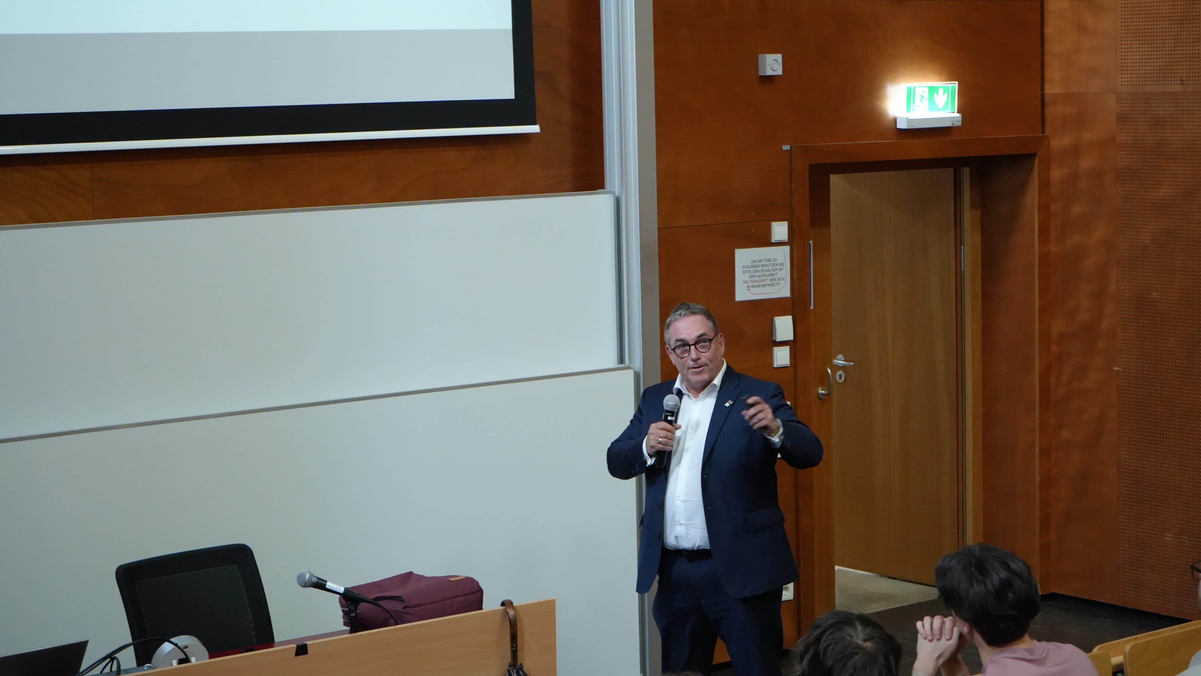 Ein Mann im Anzug steht vorne im Hörsaal, spricht in ein Mikrofon und gestikuliert dabei mit der Hand. Im Publikum sitzen mehrere Personen, und über ihm ist eine Leinwand zu sehen.