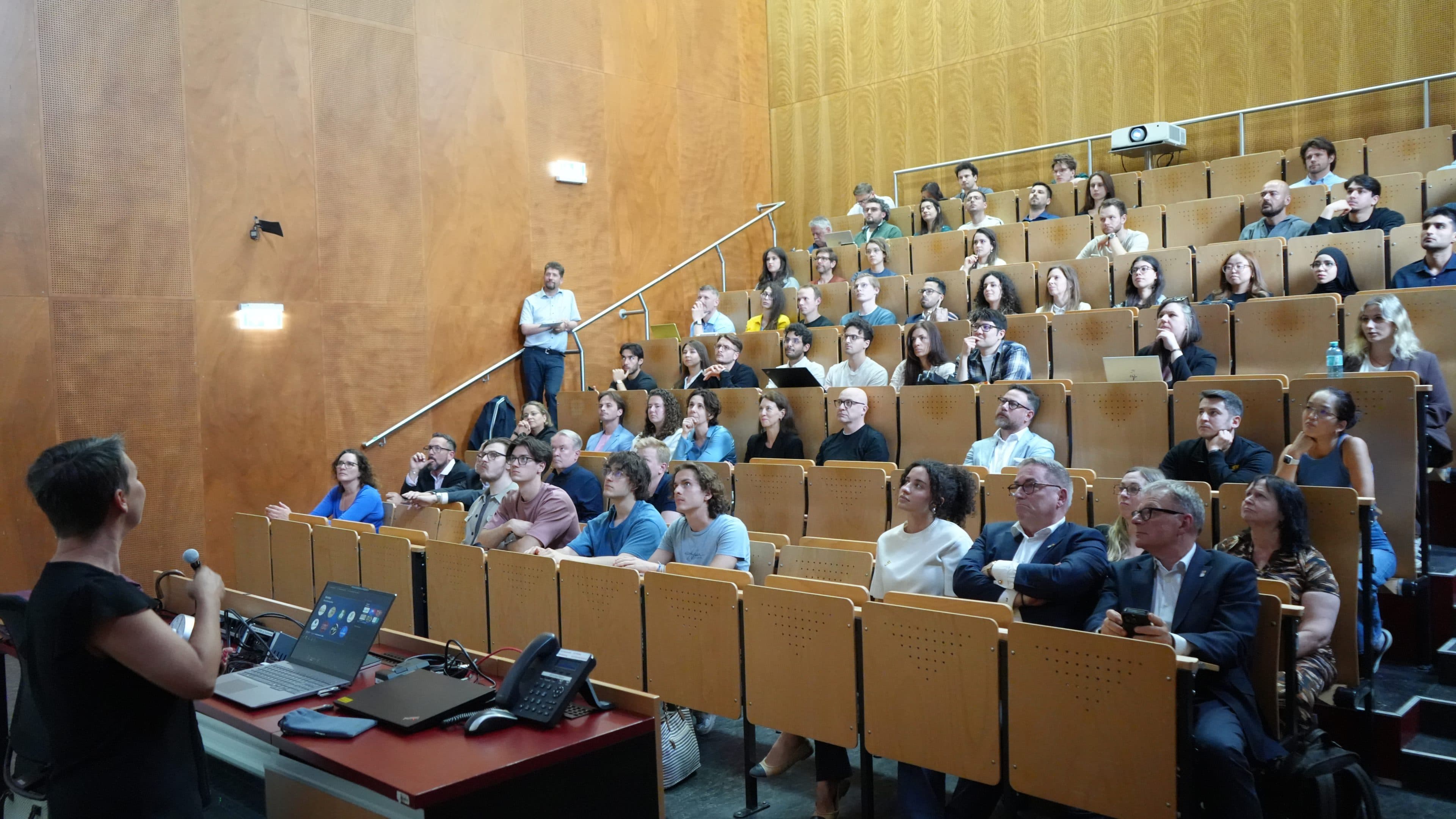 Eine Person steht auf einem Podium und hält vor einem vollbesetzten Hörsaal einen Vortrag, in dem aufgestellte Sitzreihen mit aufmerksamen Zuhörern unterschiedlichen Alters und Geschlechts in einem mit Holz vertäfelten Raum sitzen.