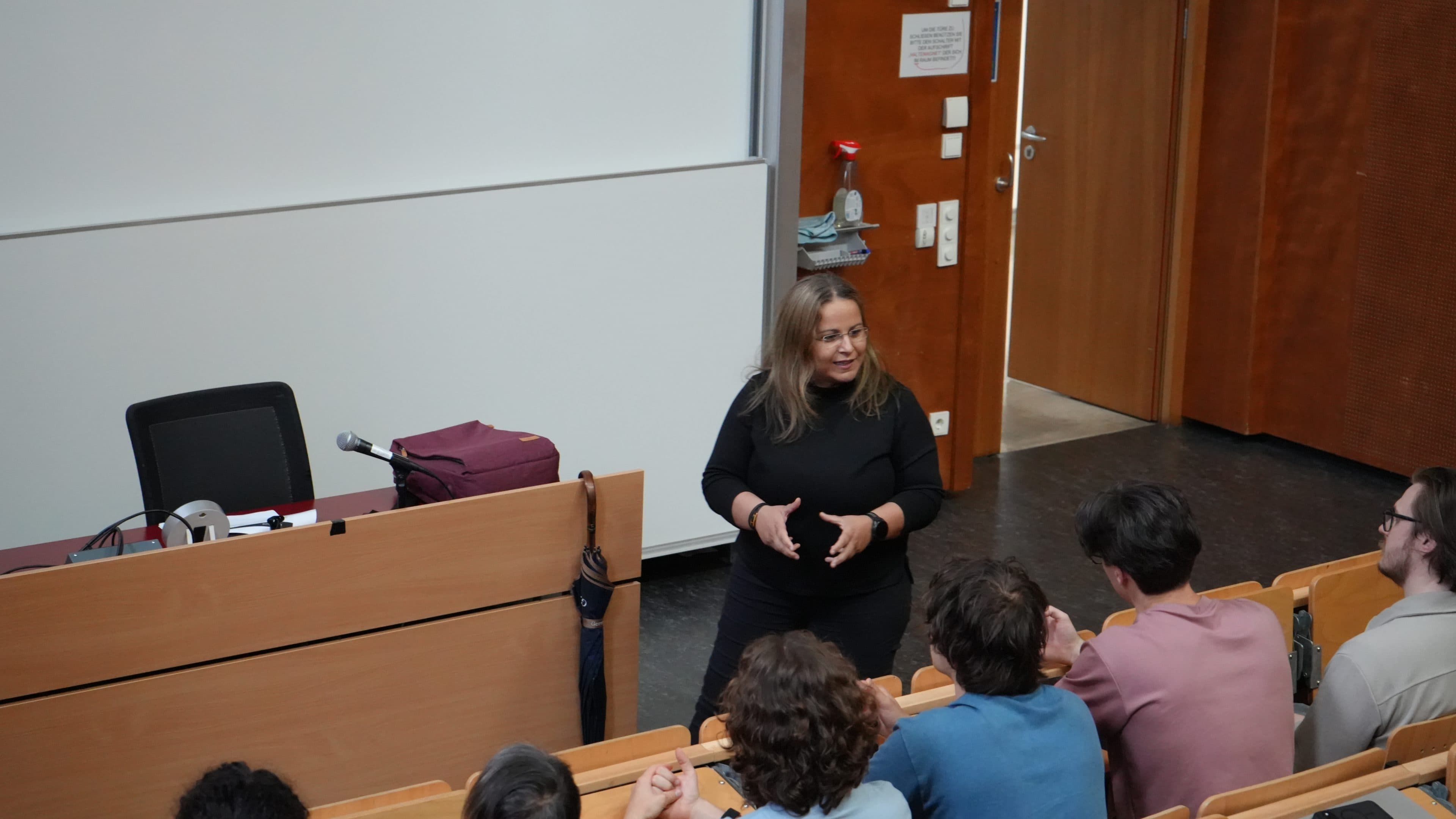 Eine Frau in schwarzer Kleidung steht vor einer Gruppe sitzender Studenten in einem Hörsaal in der Nähe eines Podiums, auf dem ein Mikrofon, eine Laptoptasche und ein geschlossener Laptop liegen. Die Studenten hören aufmerksam zu.