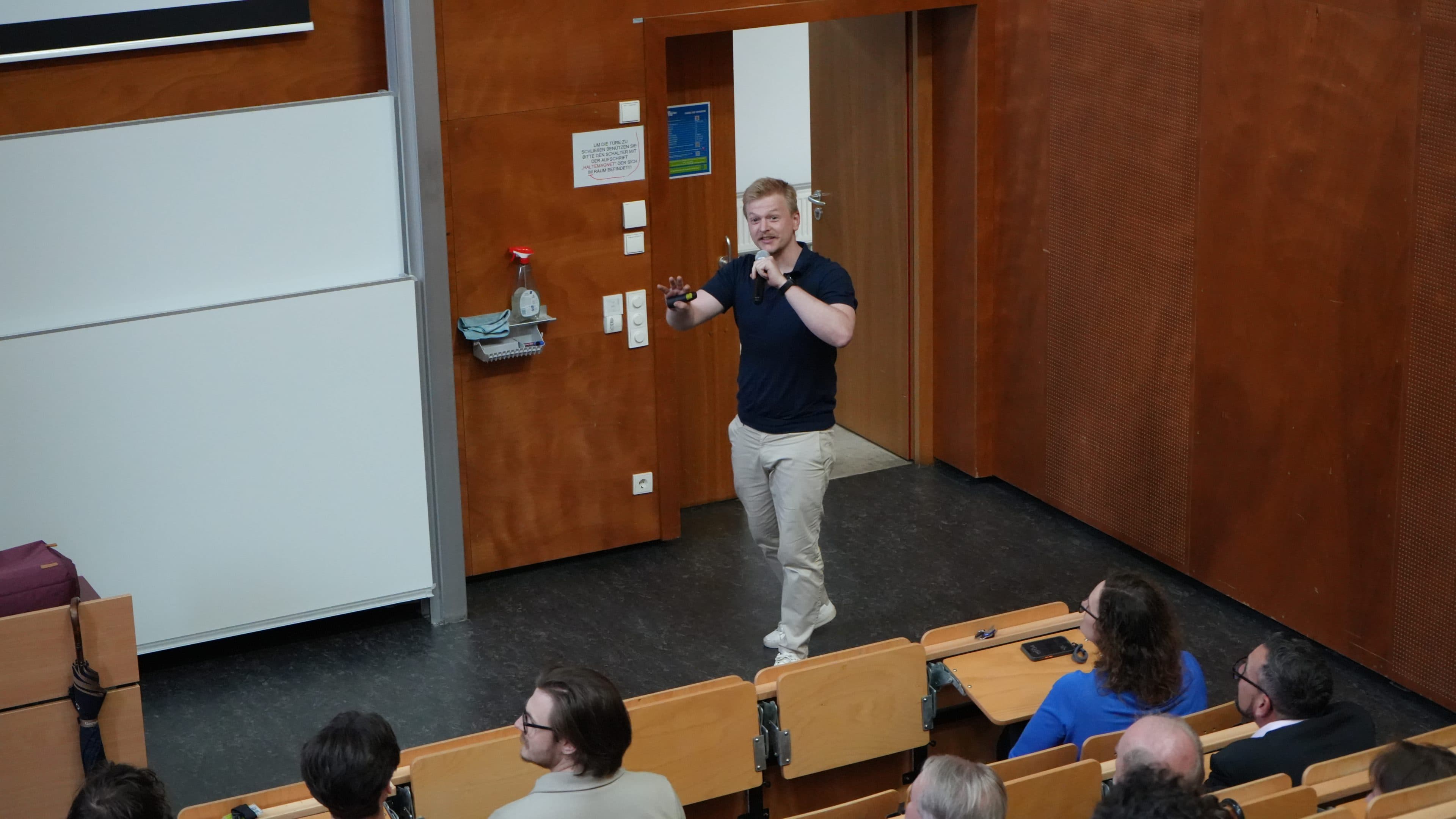 Ein Mann steht vorne im Hörsaal, spricht und gestikuliert in Richtung des Publikums, das in gestuften Reihen sitzt und aufmerksam zuhört. Der Raum hat holzvertäfelte Wände und eine Whiteboard-Tafel.