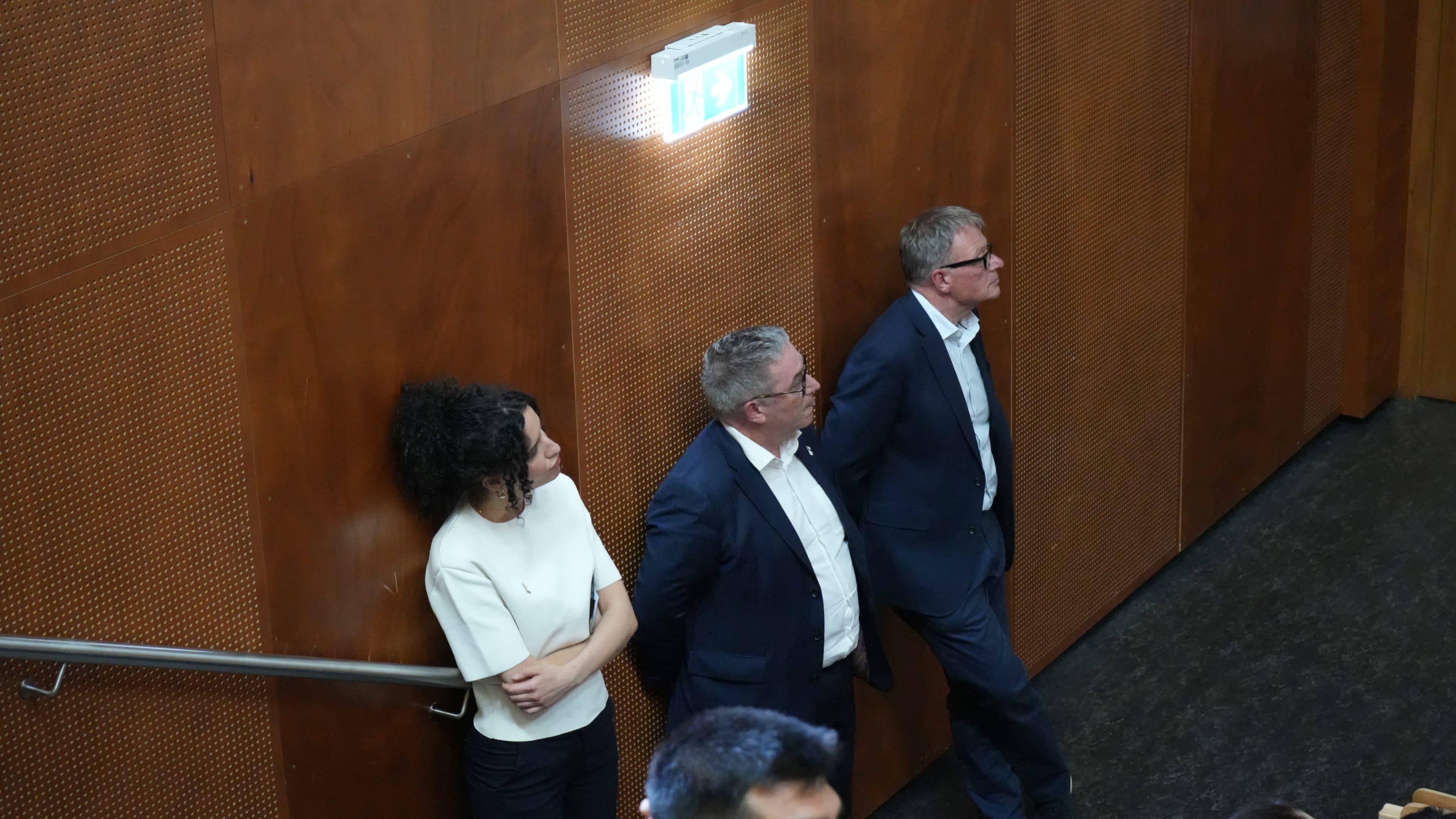 Drei Personen – zwei Männer in Anzügen und eine Frau in einem weißen Oberteil – stehen vor einer Holzwand in einem Innenraum und blicken aufmerksam nach rechts. Über ihnen befindet sich ein Notausgangsschild.