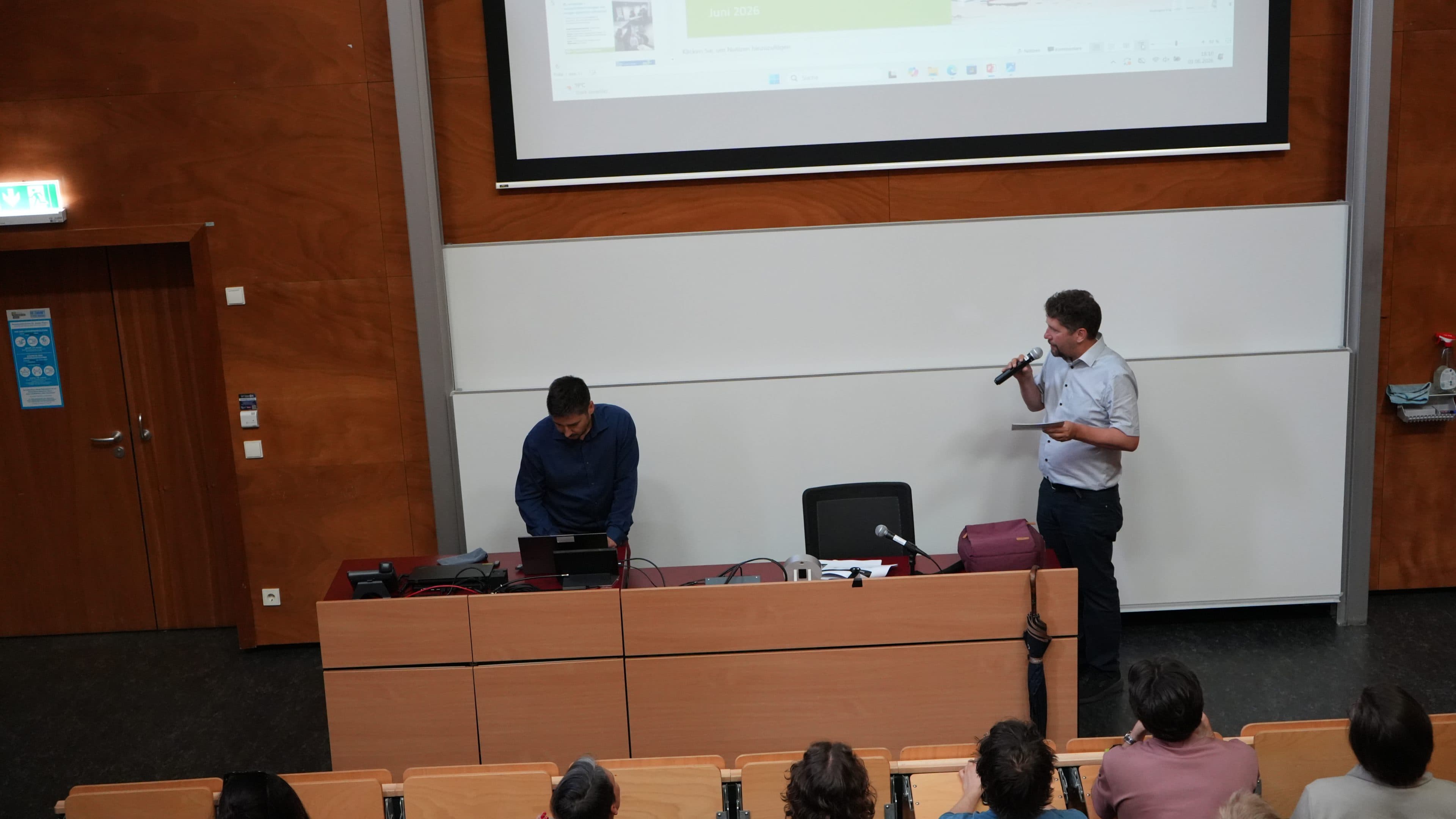 In einem Hörsaal steht ein Mann und spricht in ein Mikrofon, während neben ihm ein Mann sitzt, der an einem Laptop arbeitet; im Hintergrund sind eine Projektionswand und mehrere Zuhörer zu sehen.