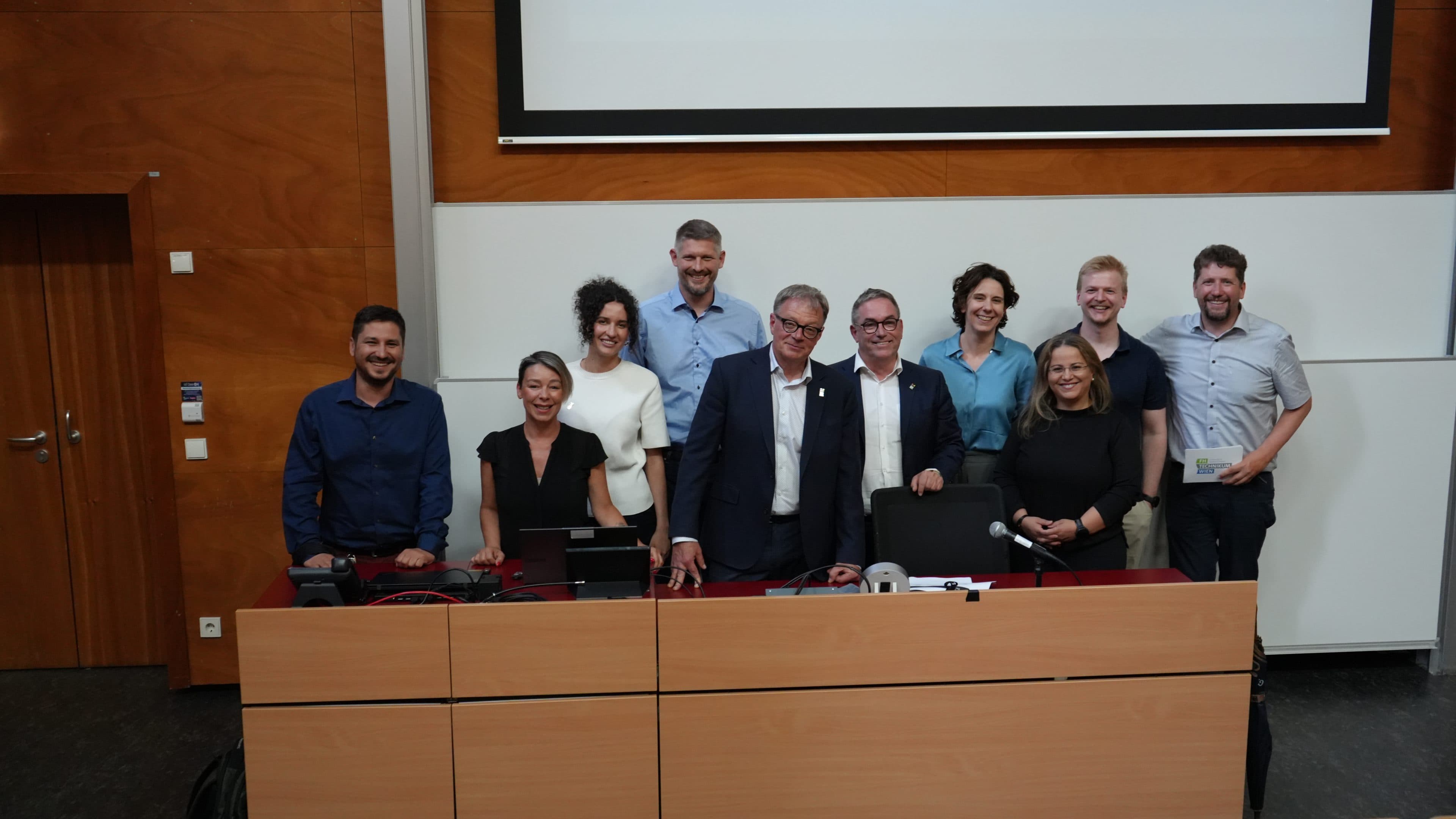 Zehn Personen stehen und sitzen gemeinsam vorne im Hörsaal und lächeln für ein Gruppenfoto vor einer großen Leinwand und Holzwänden. Einige stehen, während andere hinter einem Schreibtisch sitzen.