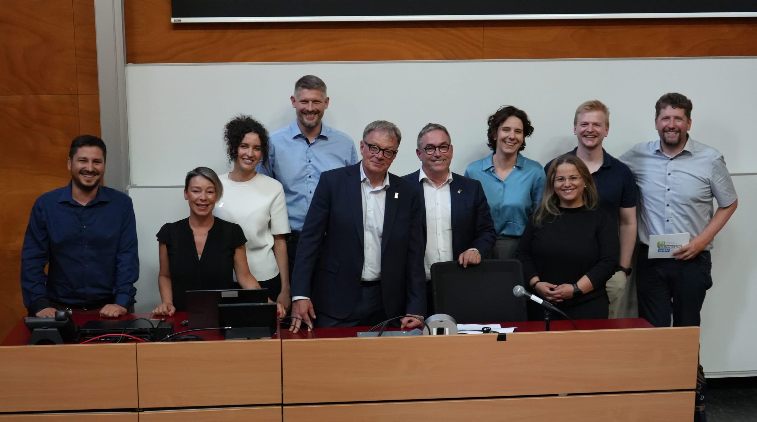 Eine Gruppe von elf Personen posiert und lächelt für ein Foto hinter einem hölzernen Podium in einem Hörsaal, hinter dem sich eine Whiteboard-Tafel und eine Projektionswand befinden.