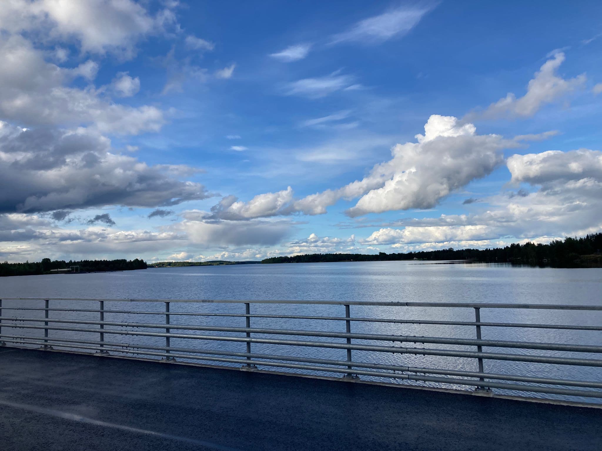 Blick von einer Brücke auf einen ruhigen See, im Vordergrund ein Metallgeländer. Der Himmel ist größtenteils blau mit vereinzelten, flauschigen weißen Wolken, und in der Ferne ist das mit Bäumen bewachsene Ufer zu sehen.