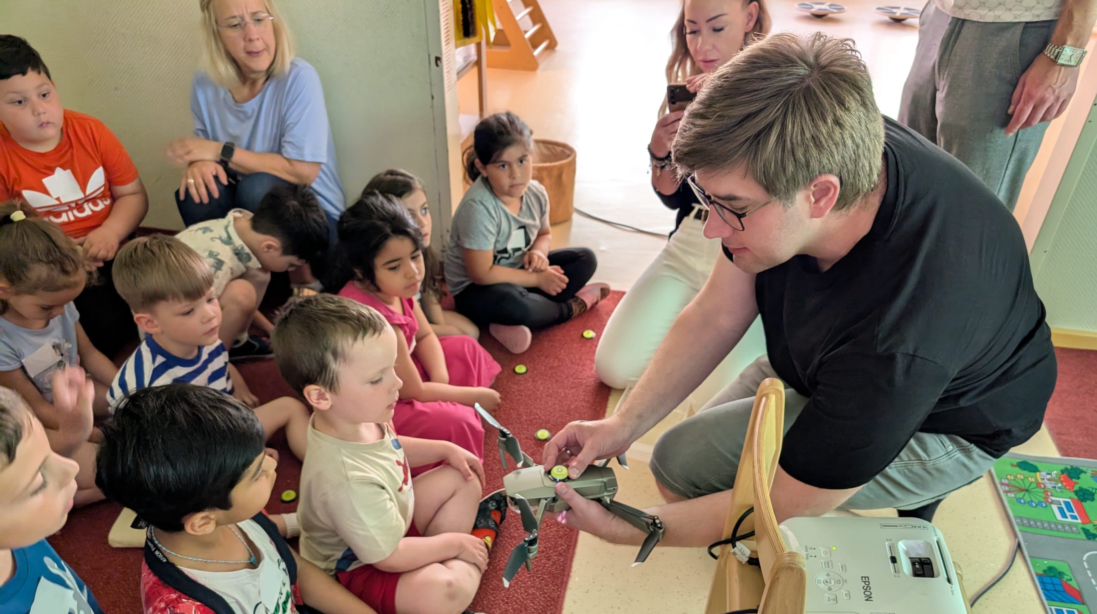 Eine Gruppe kleiner Kinder sitzt auf dem Boden und schaut zu, wie ein Erwachsener eine kleine Drohne vorführt. Einige Kinder halten gelbe Spielsteine in der Hand, und im Hintergrund beobachten Erwachsene das Geschehen. In der Nähe sind außerdem ein Projektor und eine bunte Matte zu sehen.