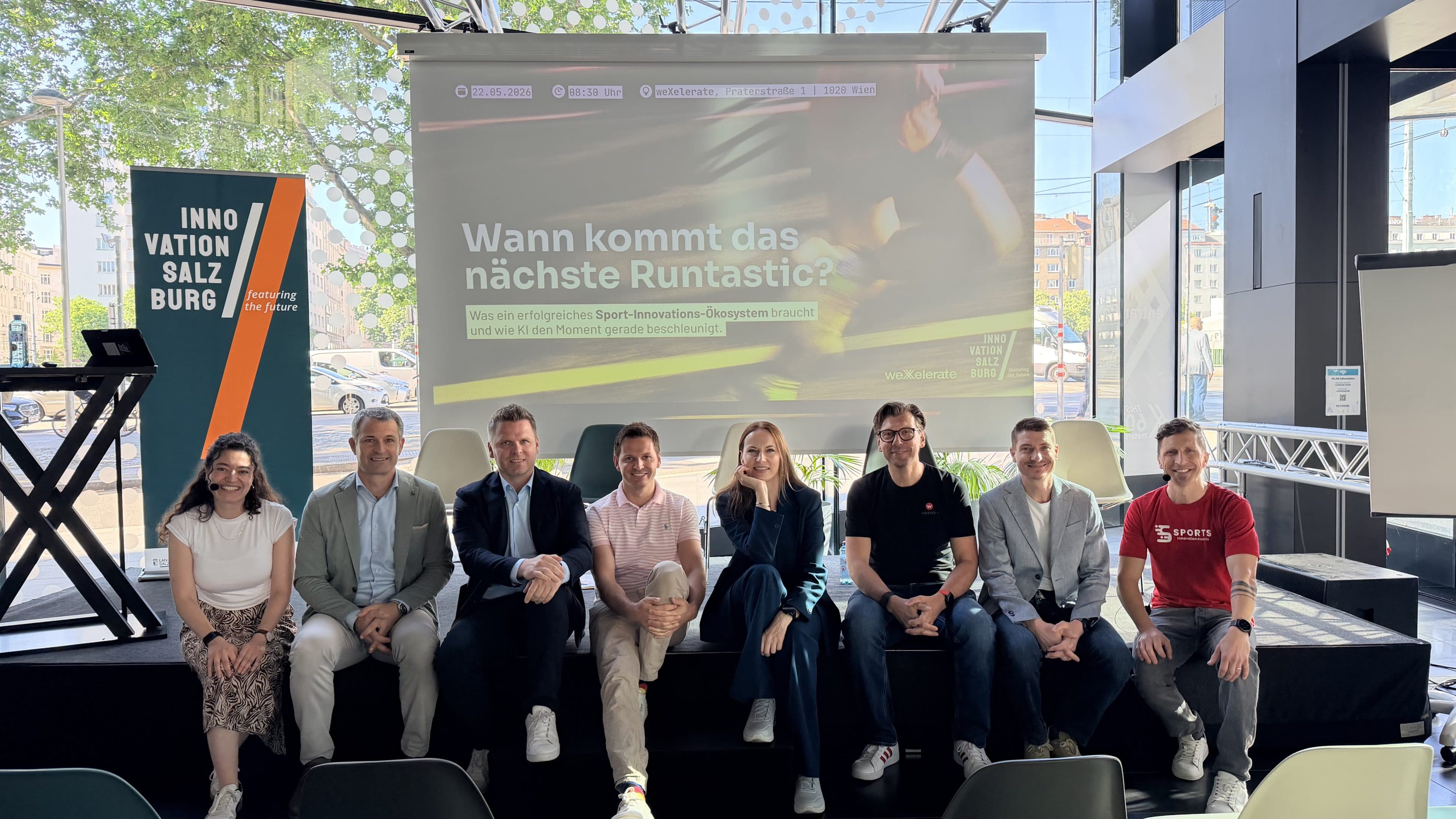 Bei einer Veranstaltung von „Innovation Salzburg“ sitzen acht Personen in einer Reihe auf der Bühne vor einer Leinwand, auf der ein deutscher Text über Runtastic projiziert wird. Helles Tageslicht strömt durch die großen Fenster hinter ihnen herein.