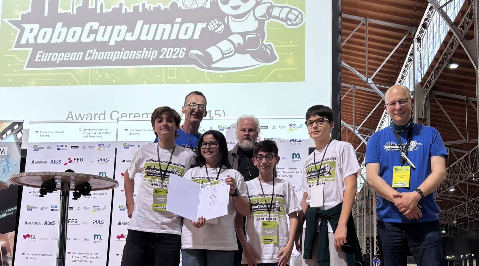 Bei der Siegerehrung der RoboCupJunior-Europameisterschaft 2026 stehen fünf Schüler und zwei Erwachsene mit einer Urkunde in der Hand auf der Bühne. Hinter ihnen ist in einer geräumigen Halle ein großes Veranstaltungsbanner zu sehen.
